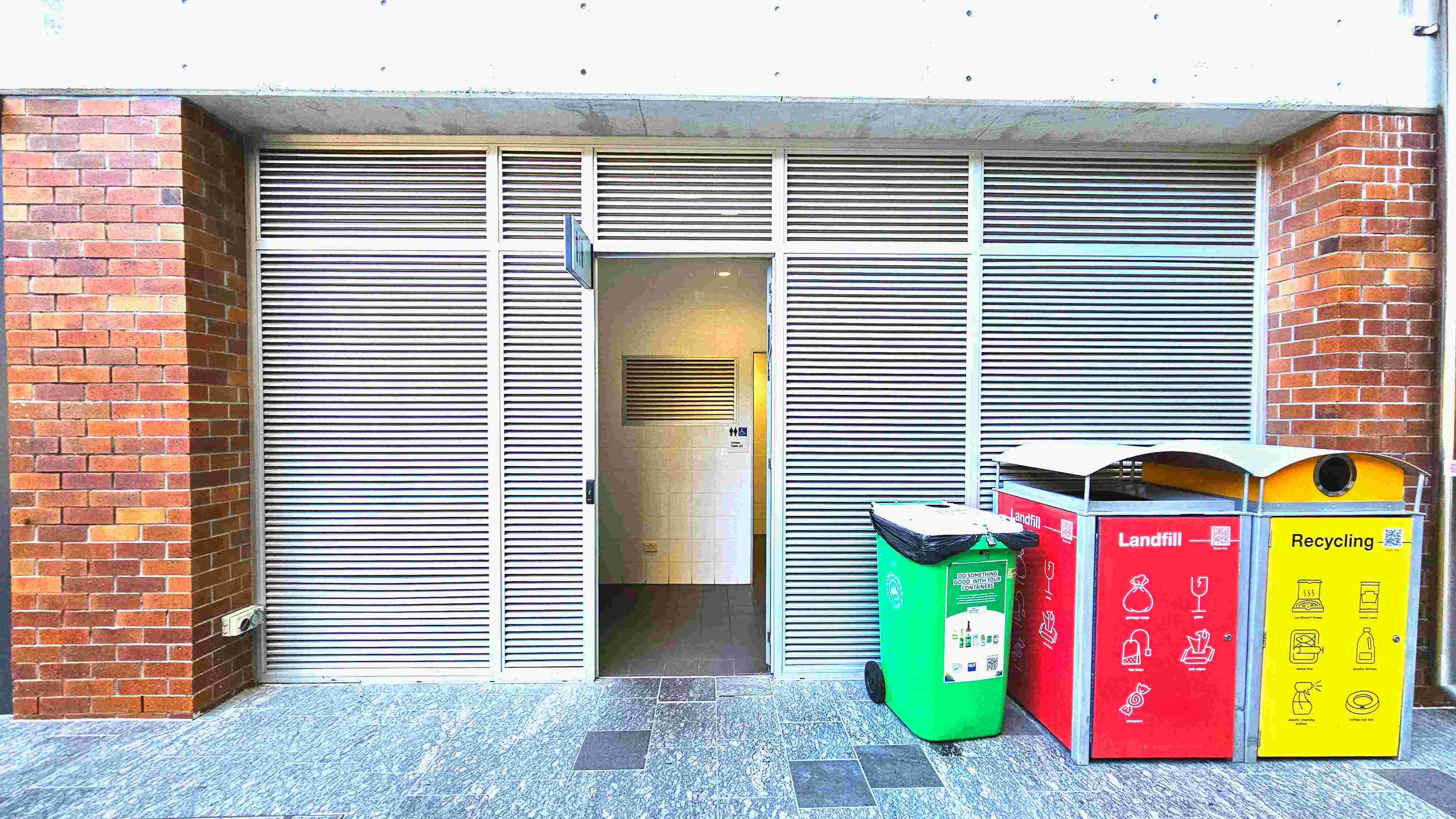 The image shows a public restroom entrance with a partially open door, set within a wall of horizontal metal slats. The entrance is flanked by red brick columns. To the right, there are three waste bins: a green one for general waste, a red one for landfill, and a yellow one for recycling. The floor is covered with grey stone tiles, some of which are darker, creating a subtle pattern. The image shows a public restroom entrance with a partially open door, set within a wall of horizontal metal slats. The entrance is flanked by red brick columns. To the right, there are three waste bins: a green one for general waste, a red one for landfill, and a yellow one for recycling. The floor is covered with grey stone tiles, some of which are darker, creating a subtle pattern.