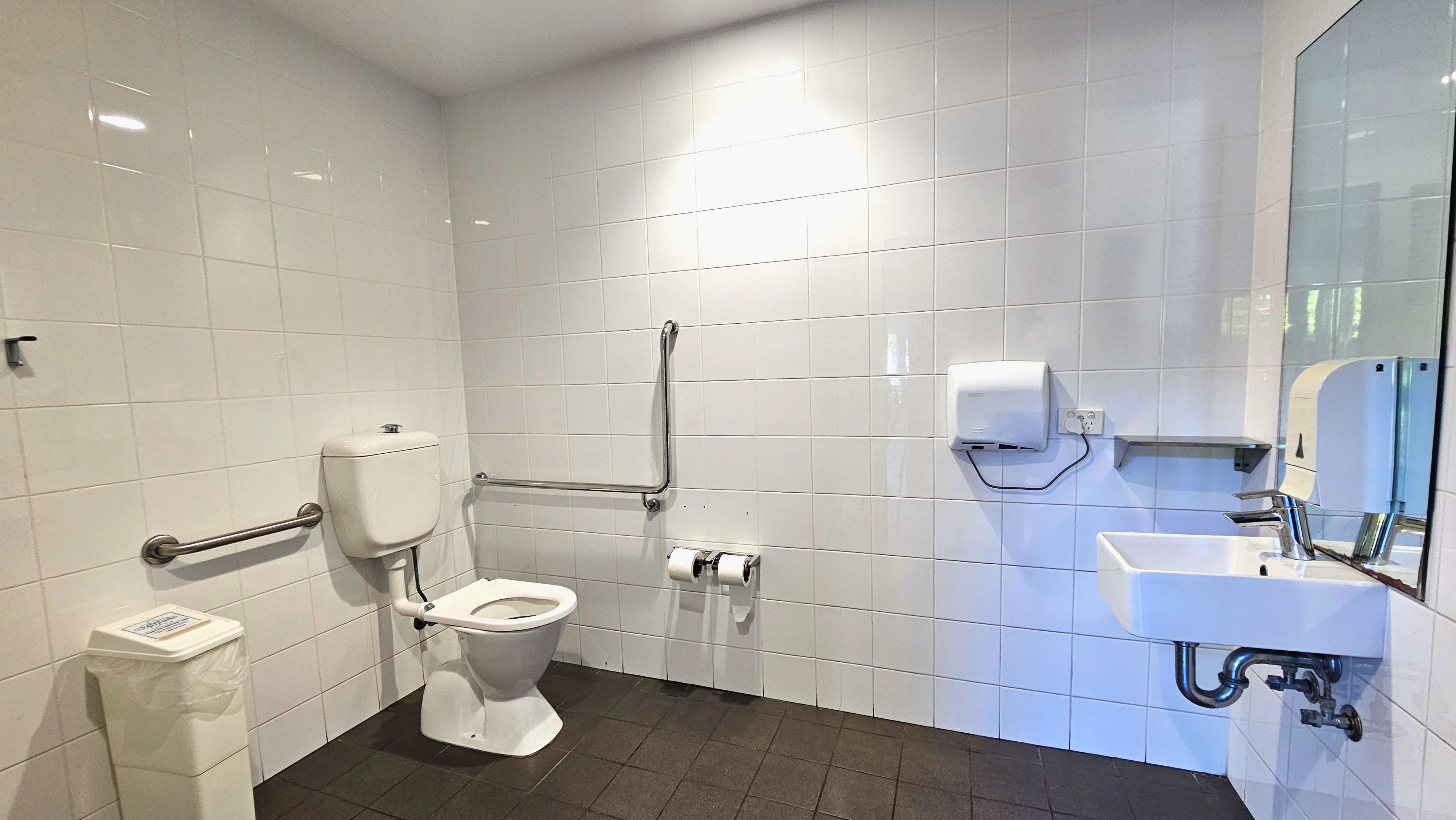 A clean, accessible bathroom with white tiled walls and a dark tiled floor. A toilet with a white cistern is positioned on the left, equipped with horizontal and vertical metal grab bars. A white bin is nearby. To the right, a wall-mounted sink with a mirror above it is visible, alongside a paper towel dispenser and a small shelf. Two toilet paper rolls are mounted on the wall between the toilet and sink. A clean, accessible bathroom with white tiled walls and a dark tiled floor. A toilet with a white cistern is positioned on the left, equipped with horizontal and vertical metal grab bars. A white bin is nearby. To the right, a wall-mounted sink with a mirror above it is visible, alongside a paper towel dispenser and a small shelf. Two toilet paper rolls are mounted on the wall between the toilet and sink.