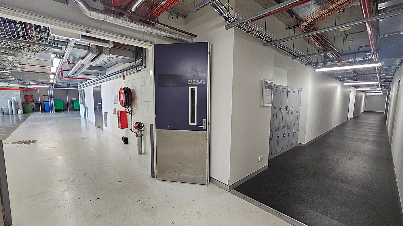A corridor with a concrete floor and ceiling, featuring exposed pipes and cables overhead. On the left, a hallway extends with fluorescent lighting and a fire hose reel on the wall. In the centre, a small room with open double doors reveals shelves inside. On the right, a large open area leads to an outdoor space with a parked vehicle and trees visible in the background. Yellow lines mark the floor near the entrance.