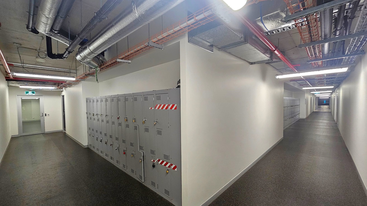 A corridor with grey lockers on the left wall, each secured with padlocks and marked with red and white stripes. The floor is dark and textured. The ceiling is exposed, showing metal pipes and cable trays. On the left, there's a grey door with an illuminated exit sign above it. The corridor extends into the distance, with fluorescent lights lining the ceiling, creating a well-lit space. The walls are plain and light-coloured.