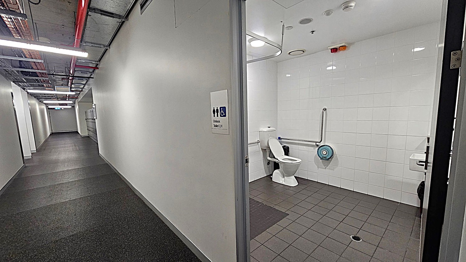 A corridor with a dark carpeted floor and a ceiling featuring exposed pipes and lighting. On the right, an open door leads to a unisex accessible toilet with white tiled walls and a grey tiled floor. Inside, there is a toilet with support rails, a toilet paper dispenser, and a small waste bin. A sign on the door indicates the toilet is unisex and accessible.