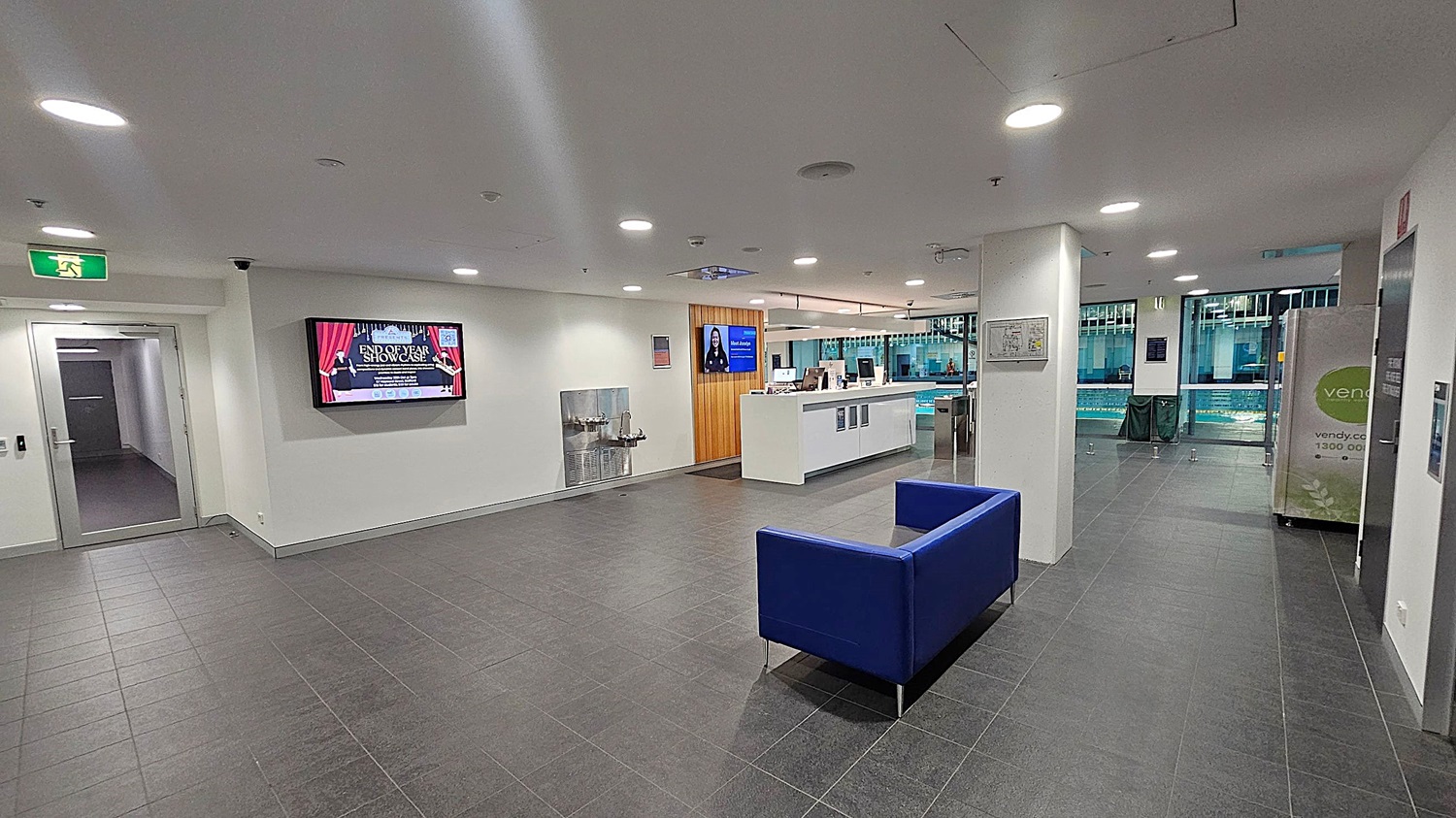 A modern lobby area with dark grey tiled flooring and a white ceiling with recessed lighting. On the left, there's a glass door and a digital screen displaying an event. In the centre, a blue sofa sits on the floor. To the right, a reception desk with computers is visible, and beyond it, large windows reveal a swimming pool area. A vending machine is positioned near the windows.