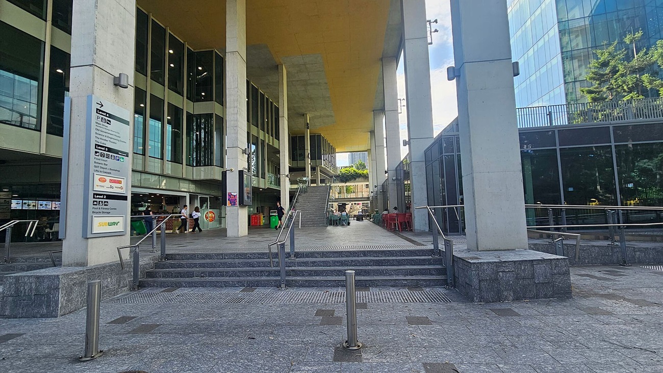 A modern building with large glass windows and tall columns. On the left, a group of people stands near the entrance, which has steps and a ramp with metal railings. The ground is paved with grey and brown tiles. On the right, a glass wall reflects the surroundings, and a cylindrical building is visible in the background. Trees are on the left side, providing some greenery.