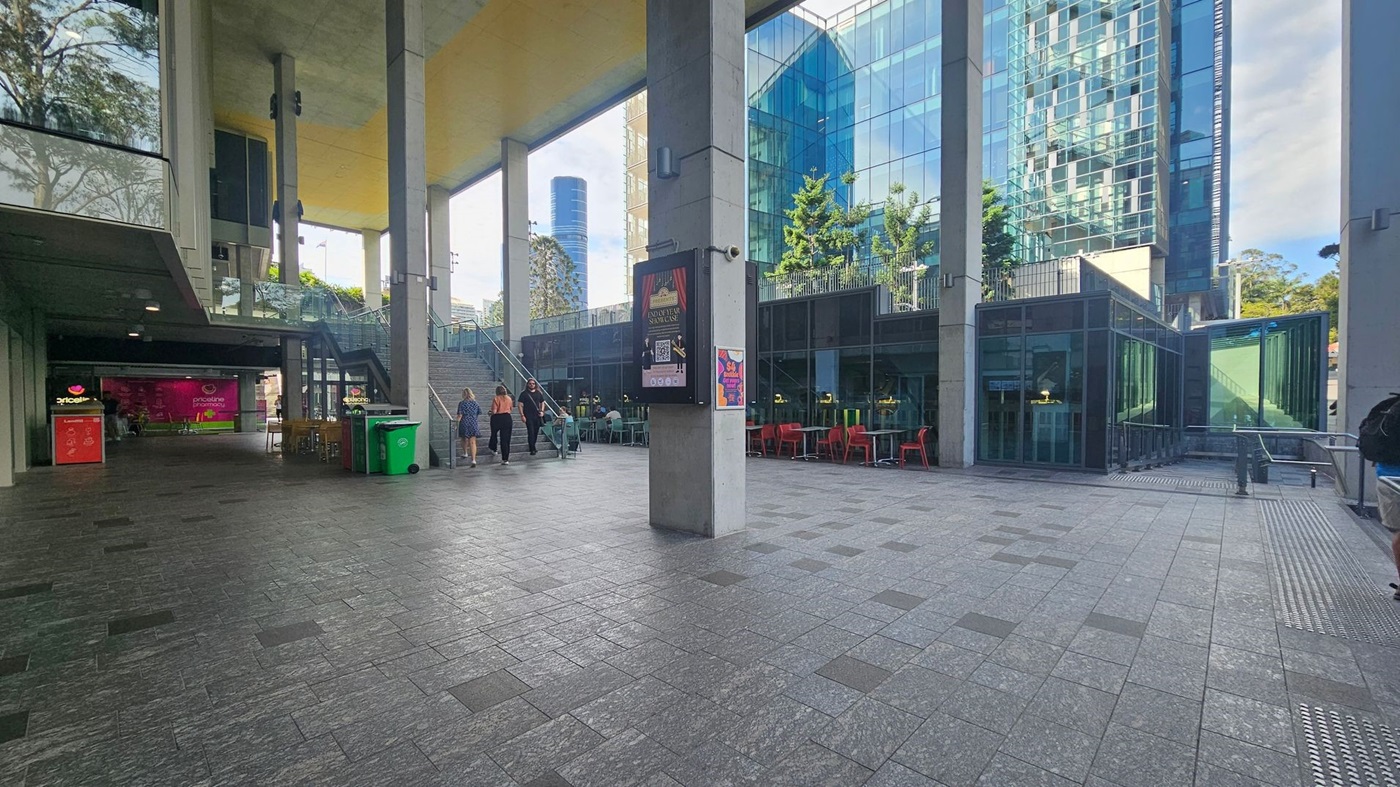 A modern urban space with a tiled floor and high ceiling. On the left, there's a covered area with a shop displaying a bright pink sign. A staircase leads up to an open area with glass railings. In the centre, a large concrete pillar has posters attached. On the right, a glass building with trees on the upper level and red chairs at outdoor tables. The background features a tall skyscraper and more greenery.