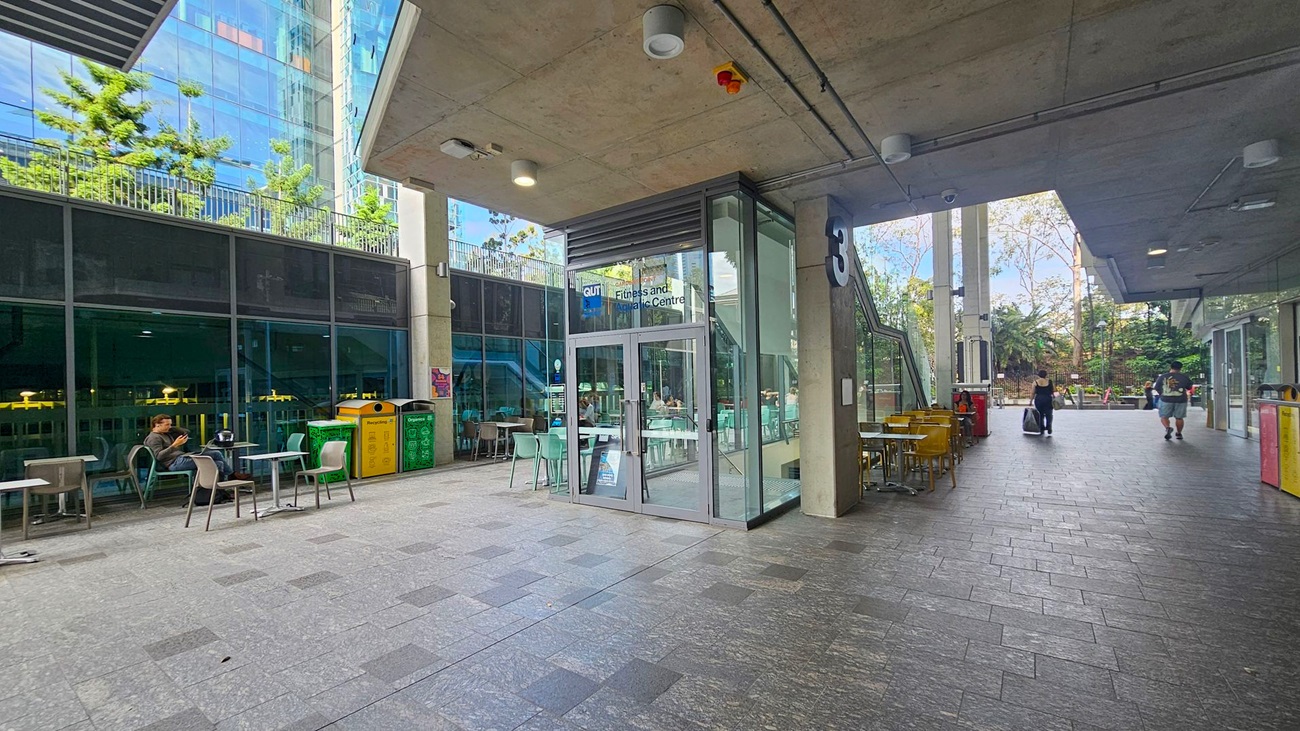 A modern outdoor area with a grey tiled floor and a concrete ceiling. On the left, there are tables and chairs, with a person seated near recycling bins. The centre features a glass entrance to a fitness and aquatic centre, with more seating inside. On the right, two people walk past more tables and bins. The background shows a glass building with reflections and greenery.