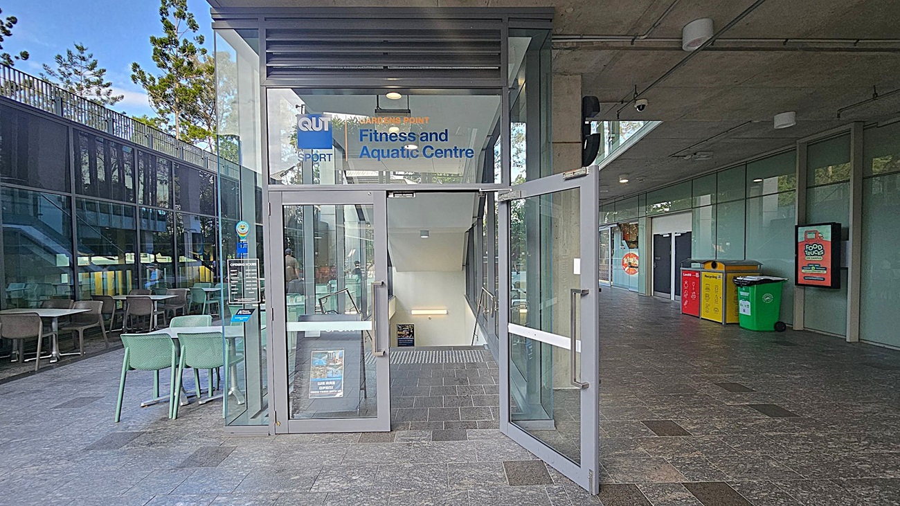 Entrance to a fitness and aquatic centre with glass doors open, revealing a staircase leading down. On the left, there are outdoor tables and chairs next to a glass wall. On the right, a row of colourful recycling bins is placed against a wall with a digital sign above. The floor is tiled in grey, and the ceiling has exposed fixtures and lights. Trees are visible through the glass.