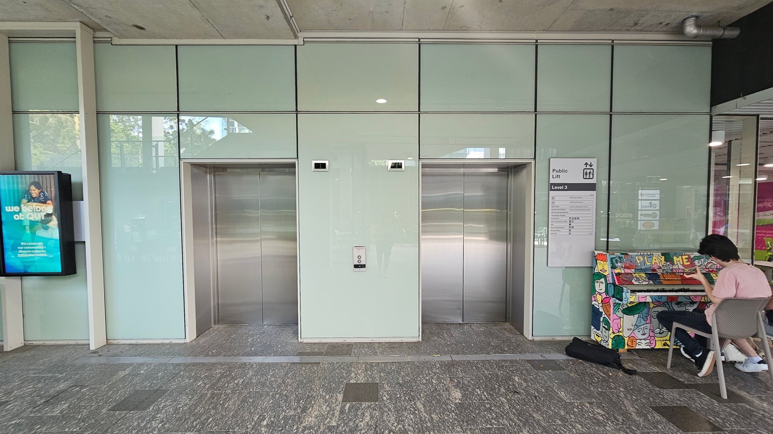 Two metallic lift doors are centred on a wall with light-coloured panels. To the left, a digital screen displays an advertisement. On the right, a person in a pink shirt sits on a chair playing a brightly painted piano with the words "PLAY ME" on it. A sign above the piano indicates "Public Lift Level 3". The floor is grey with a speckled pattern, and the ceiling is concrete with visible fixtures.