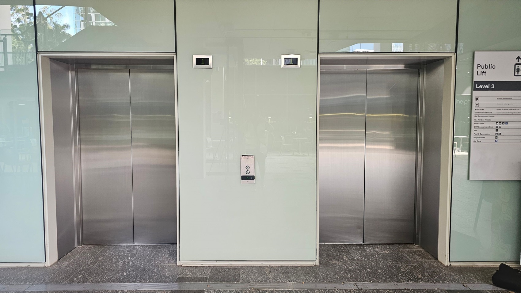 Two lift doors are set in a light-coloured wall. The left lift has a digital display above it showing the number "1". The right lift also has a digital display. Between the lifts is a control panel with buttons. On the right side of the image, a sign indicates "Public Lift" and "Level 3" with directional information. The floor is made of dark stone tiles, and there are reflections visible on the lift doors.