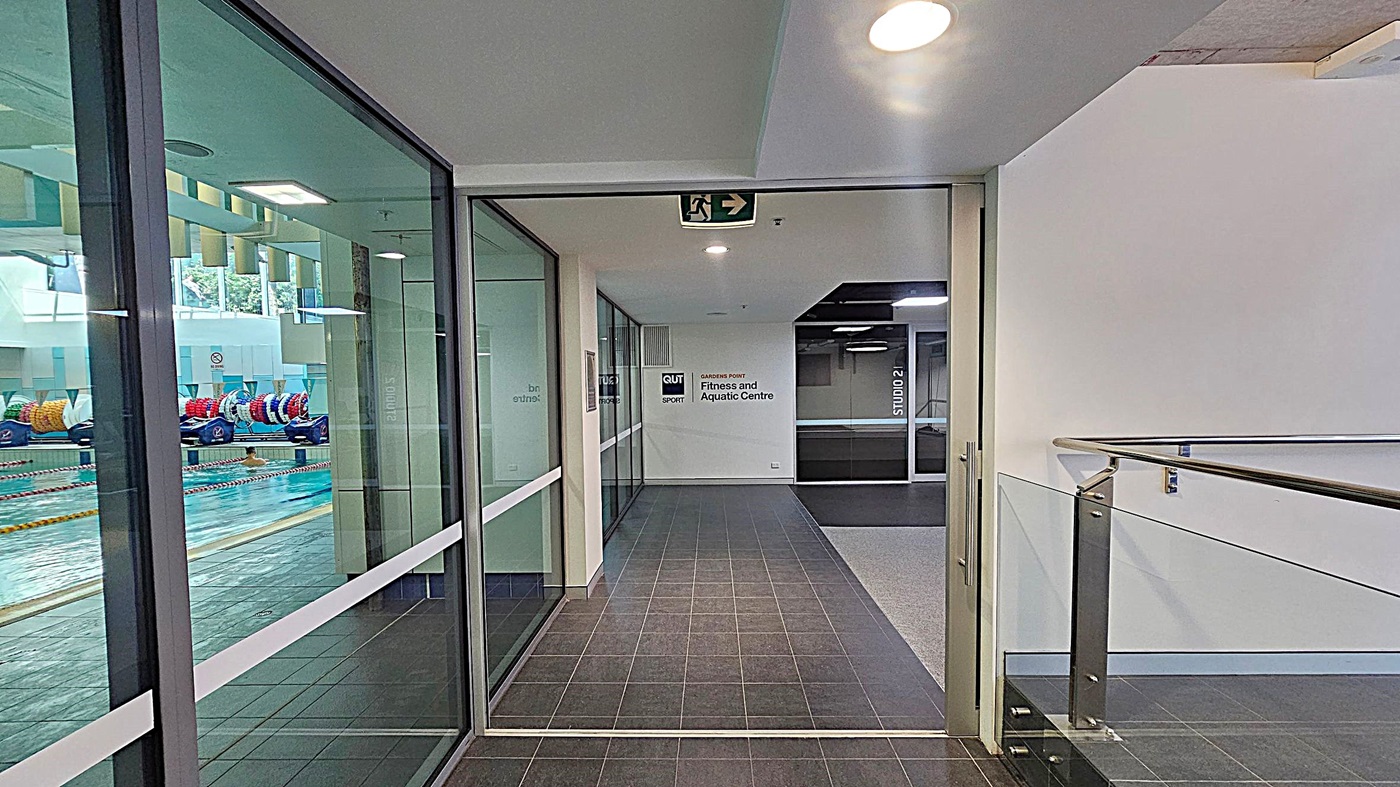 A modern indoor space with grey tiled flooring and a white ceiling with recessed lighting. On the left, there's a green bin and glass walls with signage for a fitness and aquatic centre. In the middle, a turnstile and glass barriers lead to a vending machine and doors. On the right, a reception desk and a blue chair are near two lifts. An illuminated exit sign is visible on the ceiling.