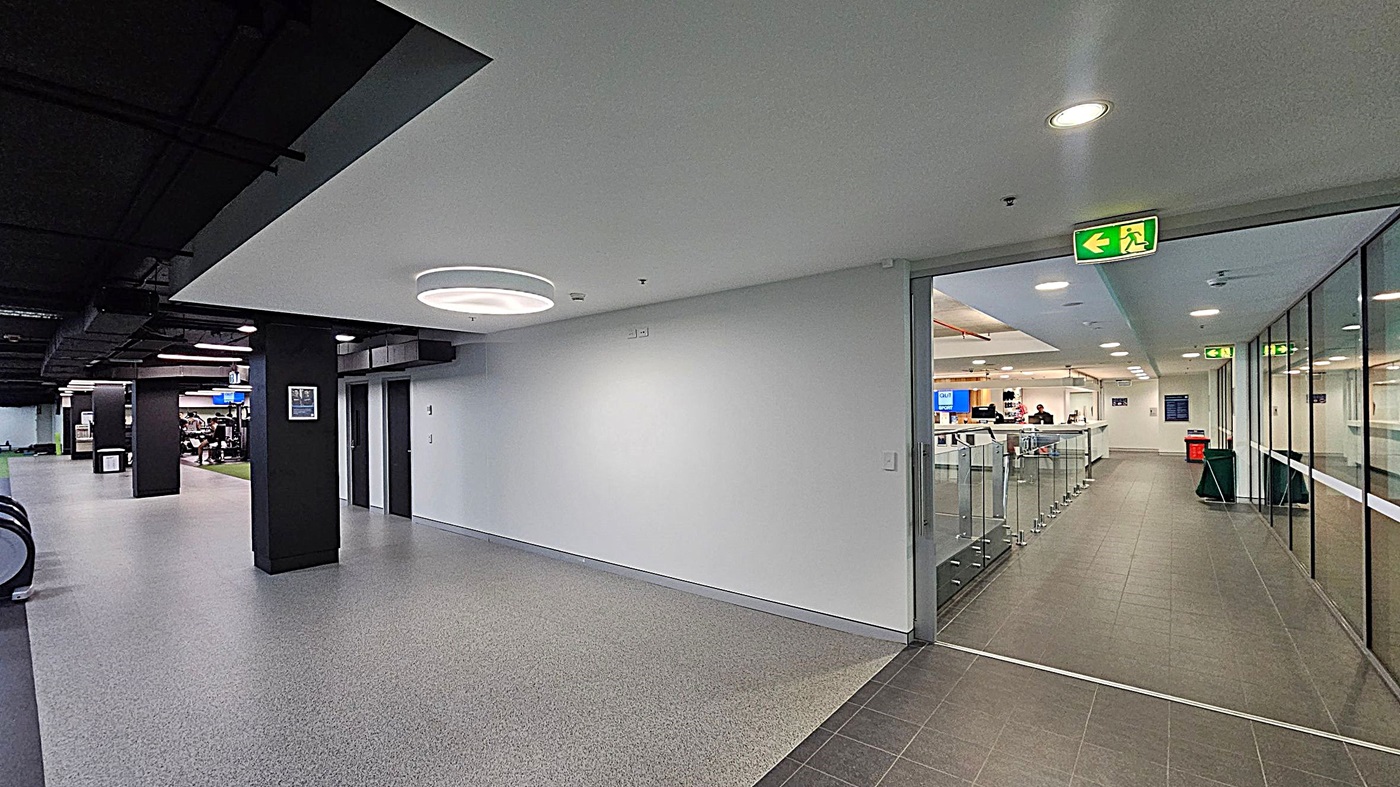 A modern indoor space with a grey speckled floor. On the left, there are escalators and a gym area with exercise equipment. The ceiling is black with recessed lighting. On the right, a glass-walled corridor leads to a reception area with a counter and people behind it. An illuminated green exit sign is above the glass doors. The walls are white, and there are black columns throughout the space.