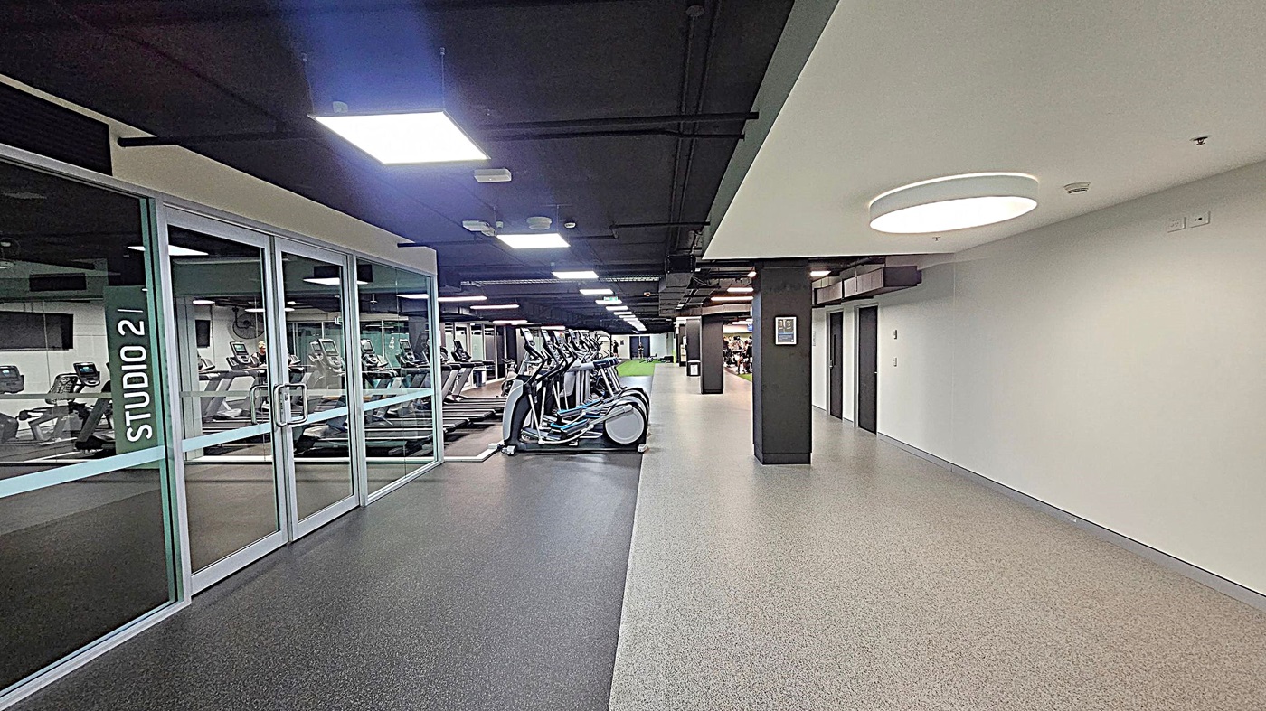 A gym interior with a corridor on the right featuring a light-coloured speckled floor and white walls. On the left, there is a glass wall with a door labelled "STUDIO 2," revealing a room with treadmills and other exercise equipment. The ceiling is dark with recessed lighting. In the centre, a row of elliptical machines is visible, and further back, more gym equipment can be seen.