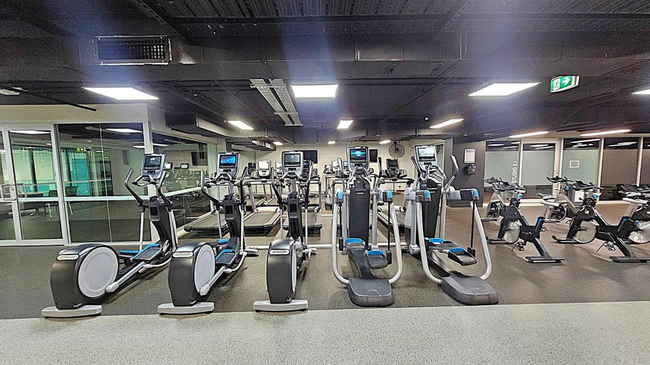 A gym with a row of elliptical machines and treadmills in the centre. To the right, there are several stationary bikes. The floor is covered with a speckled grey and green carpet. The ceiling is black with rectangular lights. On the left, there is a glass wall with a door leading to another room. An exit sign is visible on the ceiling to the right.