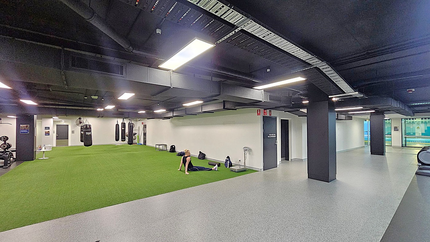 A gym interior with a large area of green artificial turf on the left, where a person is sitting on the floor. Several punching bags hang from the ceiling. On the right, there is a grey speckled floor with black pillars and a treadmill. The ceiling is black with exposed ductwork and bright rectangular lights. In the background, there are doors and a window showing an outdoor view.
