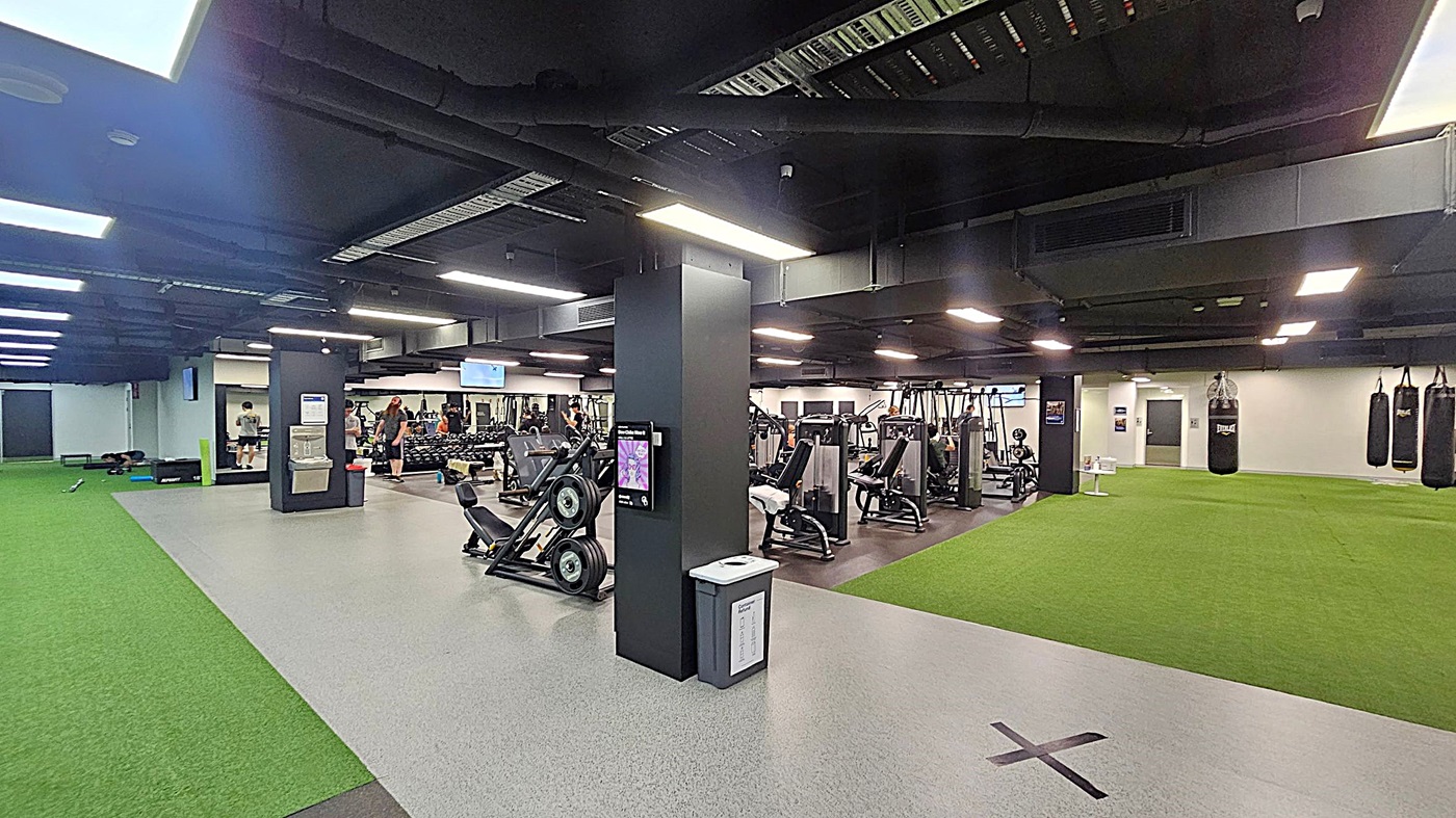 A spacious gym interior with a variety of exercise equipment. On the left, there's a green turf area and a grey floor leading to a workout zone. In the centre, several exercise machines and weights are visible, with people using them. A black pillar with a screen and a bin is in the foreground. On the right, more green turf and hanging punching bags are present. The ceiling is dark with exposed ductwork and bright overhead lights.
