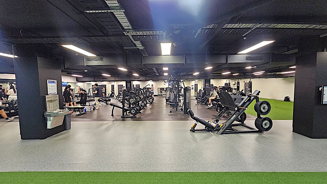 A spacious gym with various exercise equipment. On the left, there are people using weight machines and benches. In the centre, there are rows of weightlifting equipment and machines. On the right, a person is walking on a green artificial turf area. The floor is a mix of grey and green surfaces, and the ceiling is black with exposed ductwork and bright overhead lights.