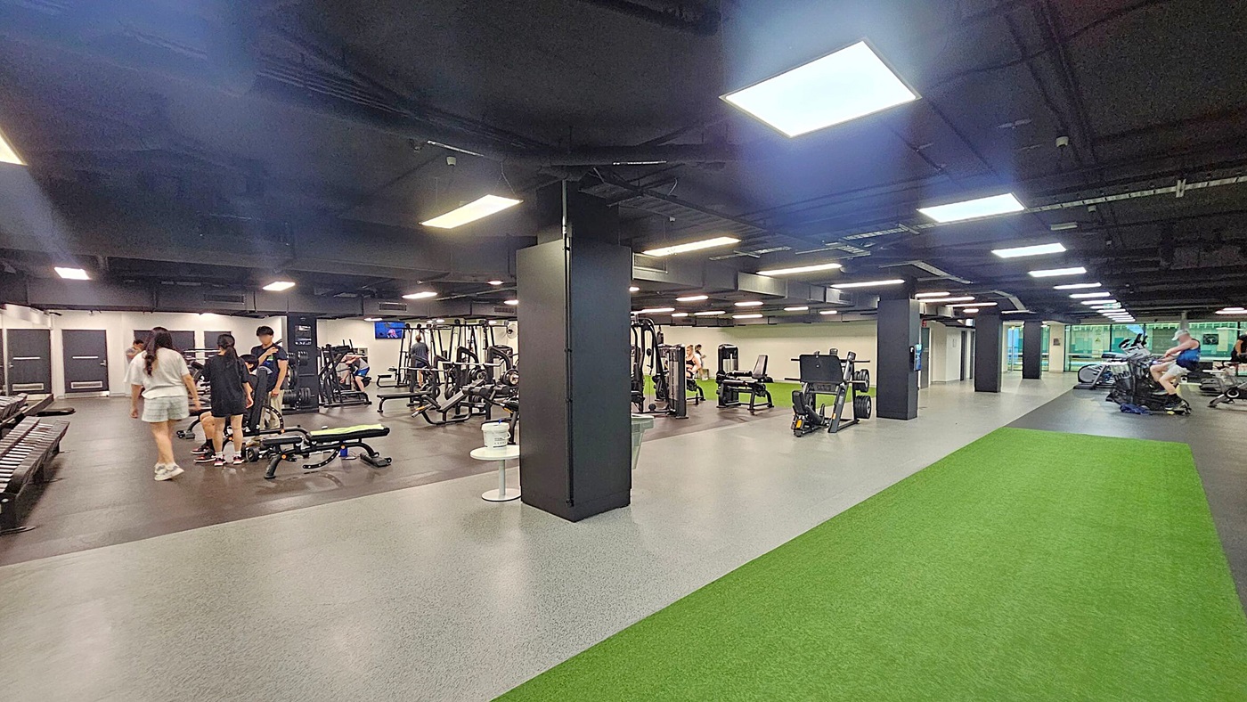 A spacious gym interior with various exercise equipment. On the left, several people are gathered near weight benches and machines. In the centre, there are more weight machines and a black pillar. On the right, a green turf strip runs along the floor, with cardio machines like stationary bikes nearby. The ceiling is dark with bright rectangular lights, and the floor is a mix of grey and green surfaces.