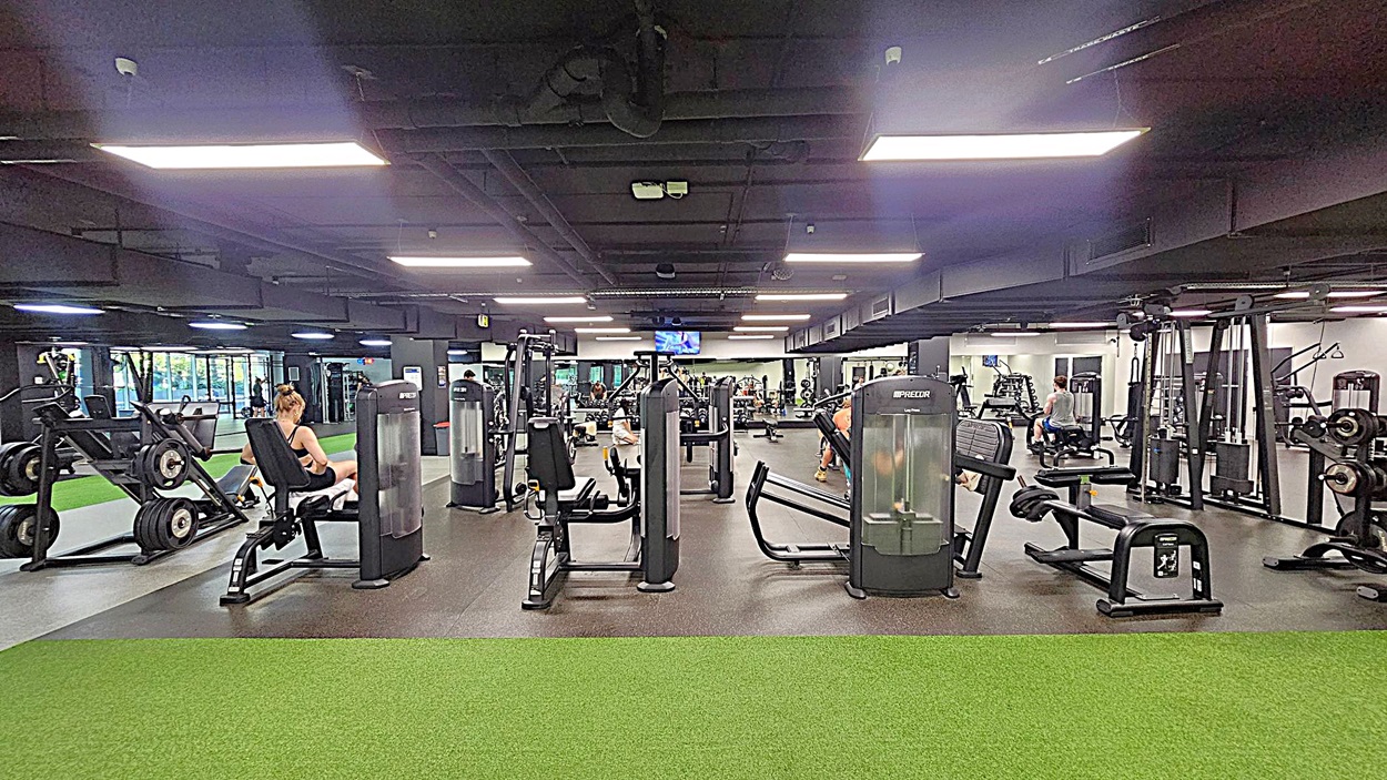 A spacious gym with various exercise machines and equipment. In the foreground, there's a green artificial turf area. The middle section features several workout machines, including leg presses and rowing machines. People are using the equipment, with one person seated on a machine to the left. The background shows more gym equipment and a wall-mounted TV. The ceiling is dark with bright rectangular lights.