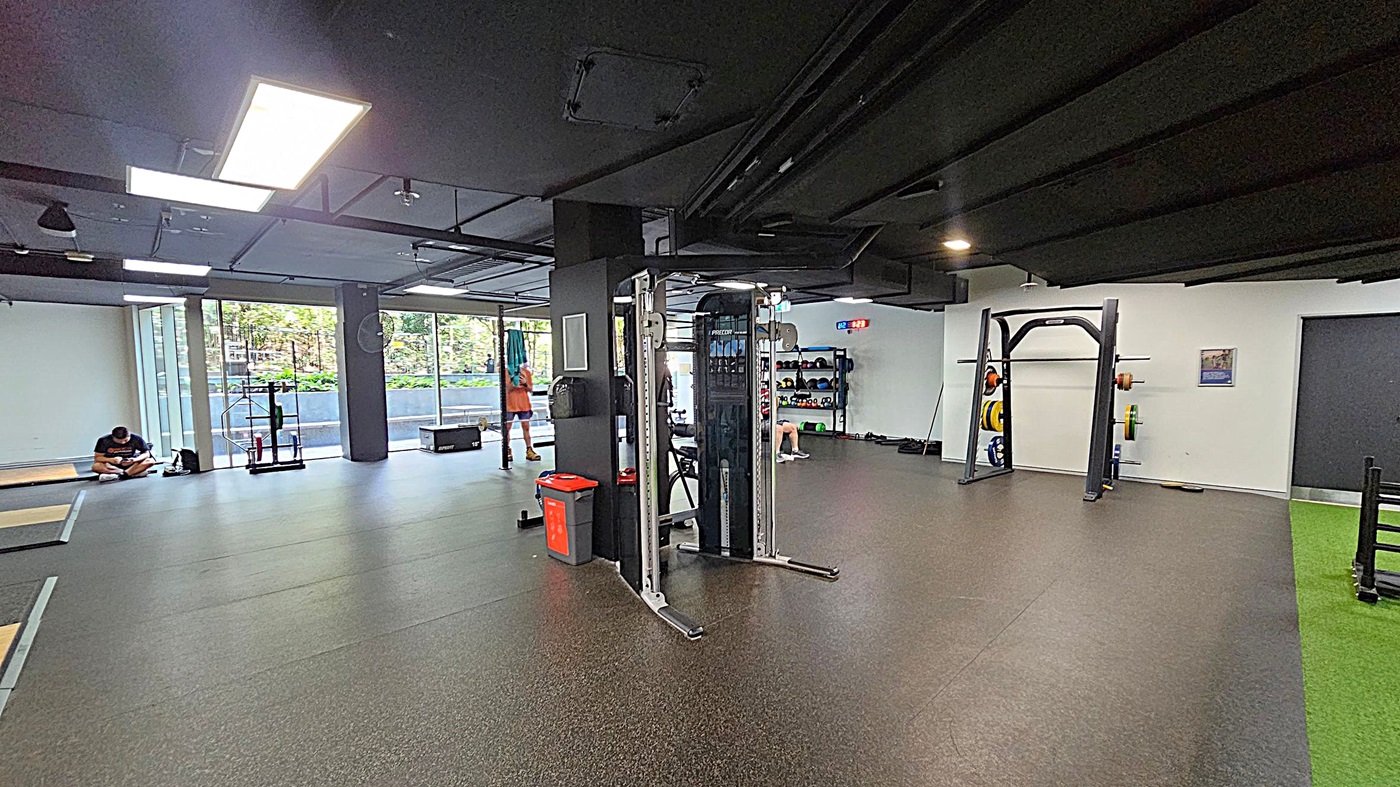 A spacious gym with a dark ceiling and rubber flooring. On the left, a person sits on the floor near large windows, with gym equipment nearby. In the centre, a multi-functional exercise machine and a red bin are visible. On the right, there is a squat rack with weights and a green turf area. The back wall has shelves with kettlebells and other equipment. Bright overhead lights illuminate the space.
