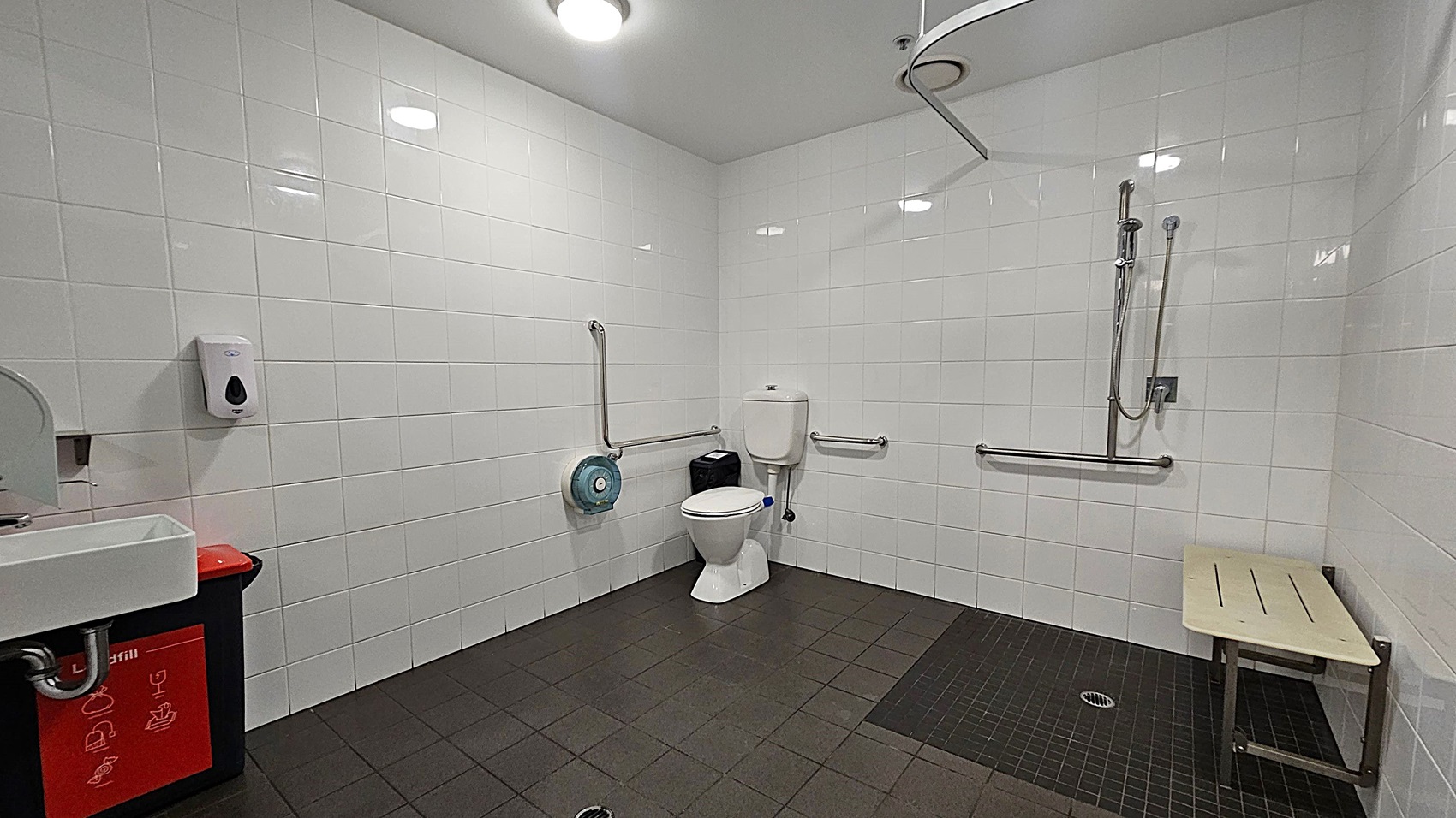 A spacious, accessible bathroom with white tiled walls and a dark tiled floor. On the left, there's a small white sink with a soap dispenser above it and a red bin below. In the centre, a toilet with support rails on both sides and a toilet paper dispenser nearby. On the right, a shower area with a handheld showerhead and a foldable wooden bench. The ceiling is white with recessed lighting.
