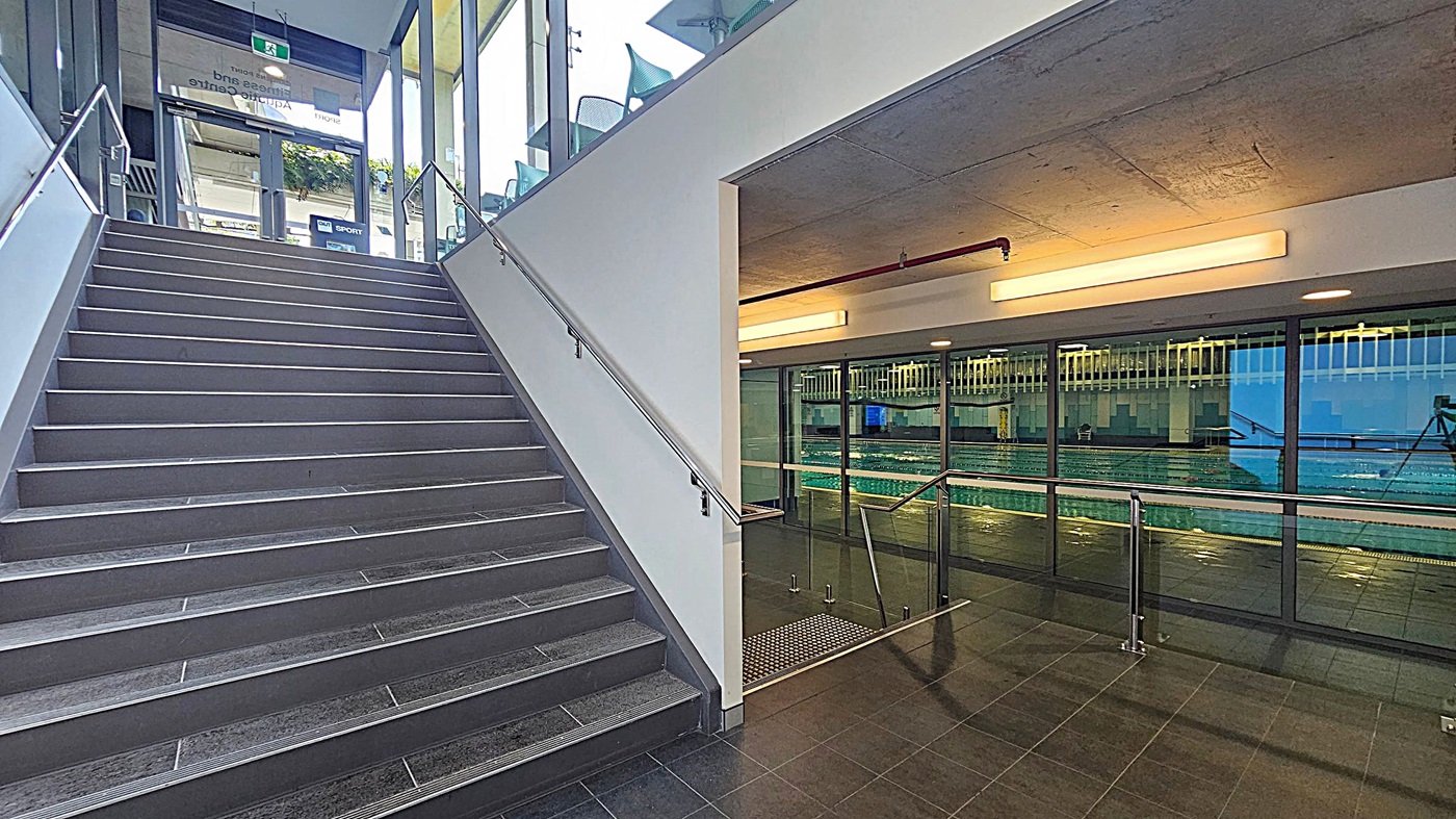 A modern indoor space with a staircase on the left, featuring grey steps and metal railings. To the right, there is a glass wall with a view of an indoor swimming pool. The ceiling is concrete with long, rectangular lights. The floor is tiled in dark grey. The area is well-lit, and there are glass doors at the top of the stairs leading outside.