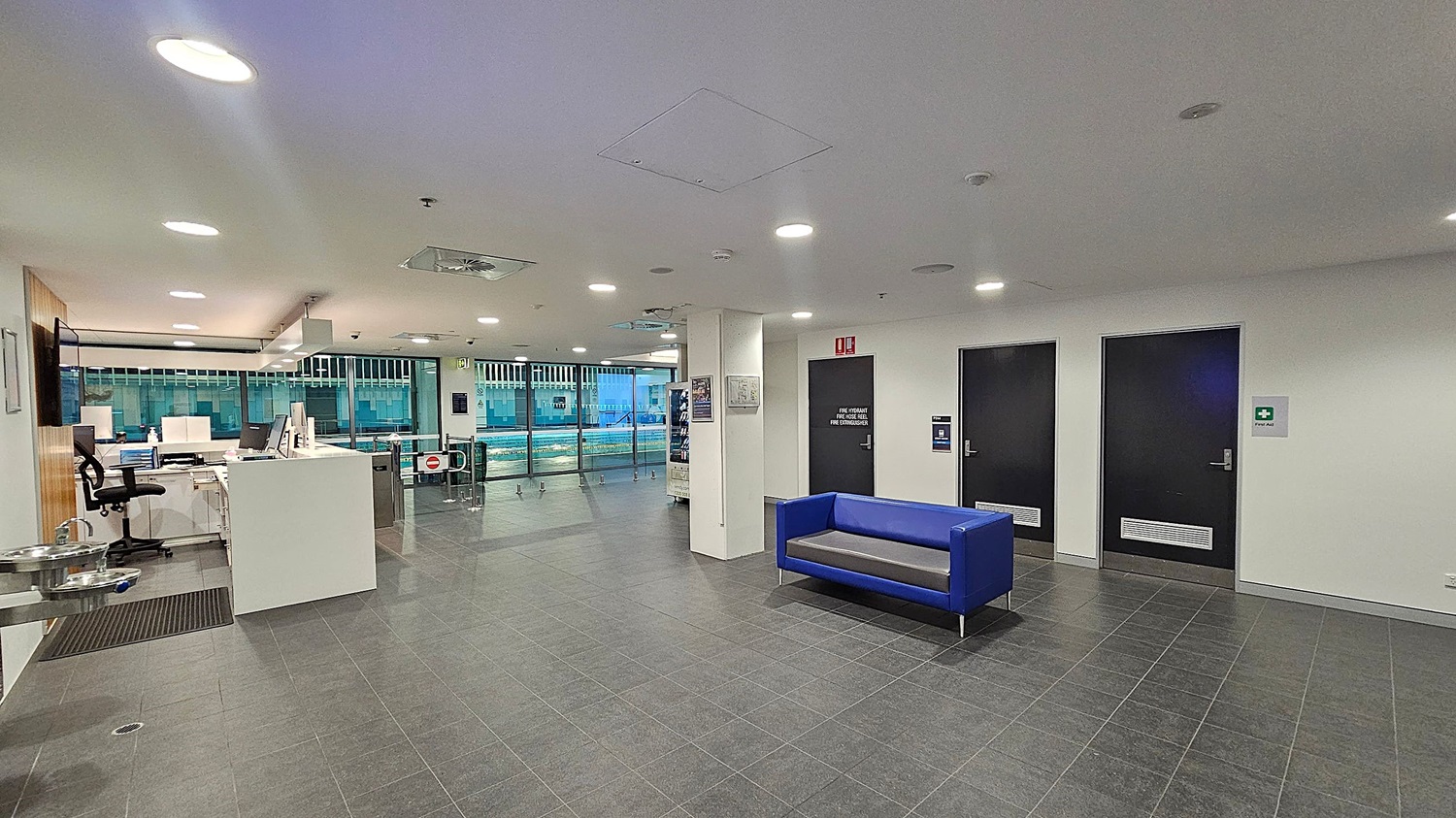 A modern indoor space with a grey tiled floor and white ceiling featuring recessed lighting. On the right, there are two black doors with signs, and a blue sofa against the wall. On the left, a reception area with a white desk, office chair, and computer equipment. A water fountain is mounted on the wall. In the background, large windows reveal a view of a swimming pool area with lanes and water.