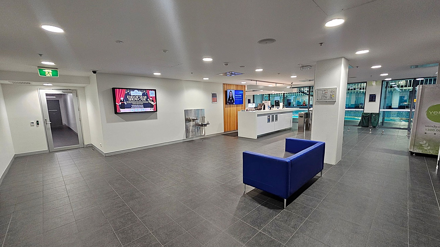 A modern indoor space with a dark tiled floor and white ceiling with recessed lighting. On the left, there's a glass door beneath an exit sign and a wall-mounted screen displaying an event. In the centre, a water fountain is visible. To the right, a reception desk with computers and another screen is present. A blue sofa is in the foreground. Large windows in the background reveal a swimming pool area. A pillar with a map and a vending machine are also on the right.