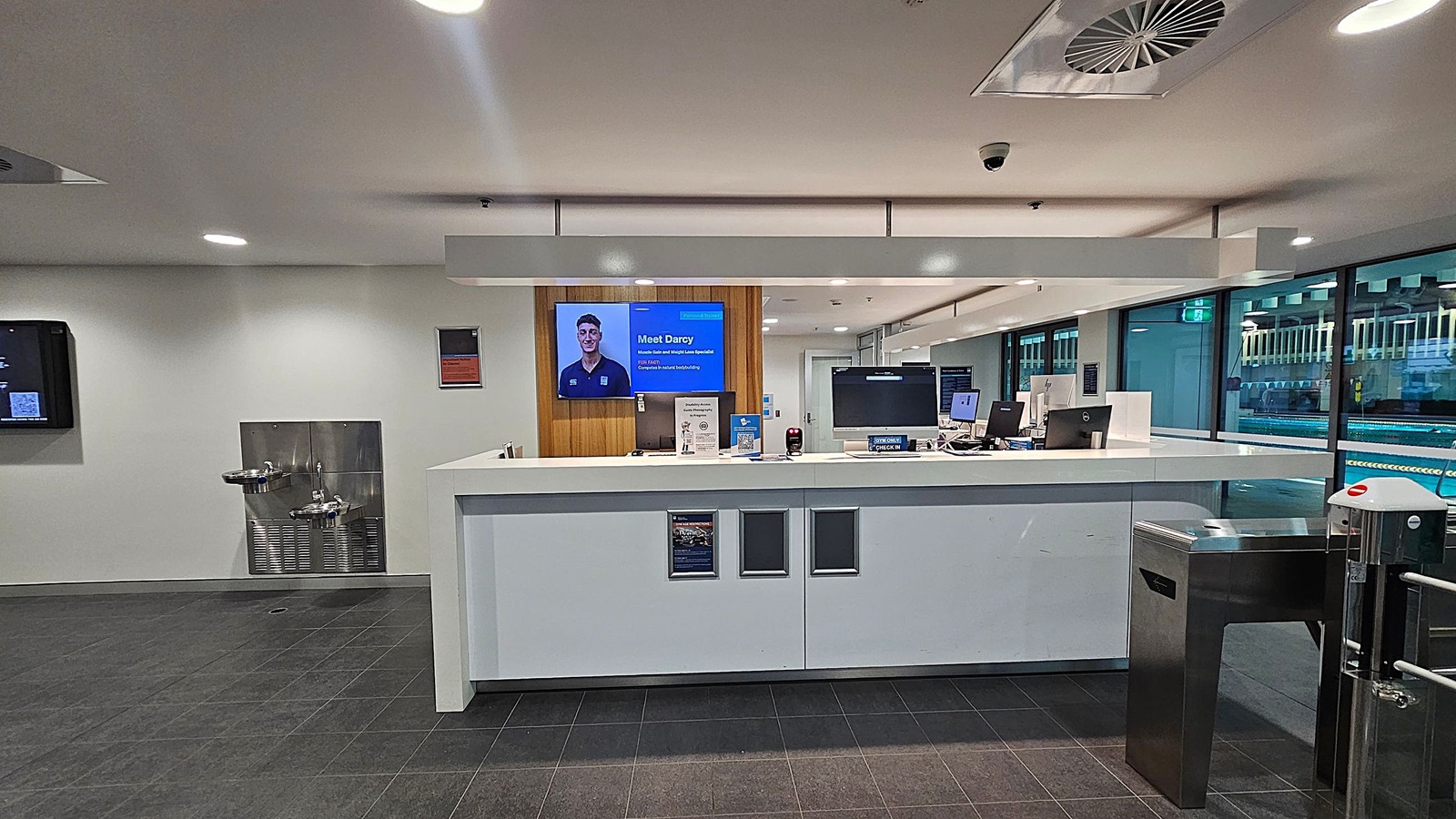 A modern reception area with a white counter in the centre, featuring computer monitors and a small sign. Behind the counter, a digital screen displays a person’s image and text. To the left, a wall-mounted water fountain and a digital display are visible. On the right, a turnstile gate is present. The floor is dark grey tile, and the ceiling is white with recessed lighting and a ventilation unit. Large windows on the right show a glimpse of a swimming pool area.