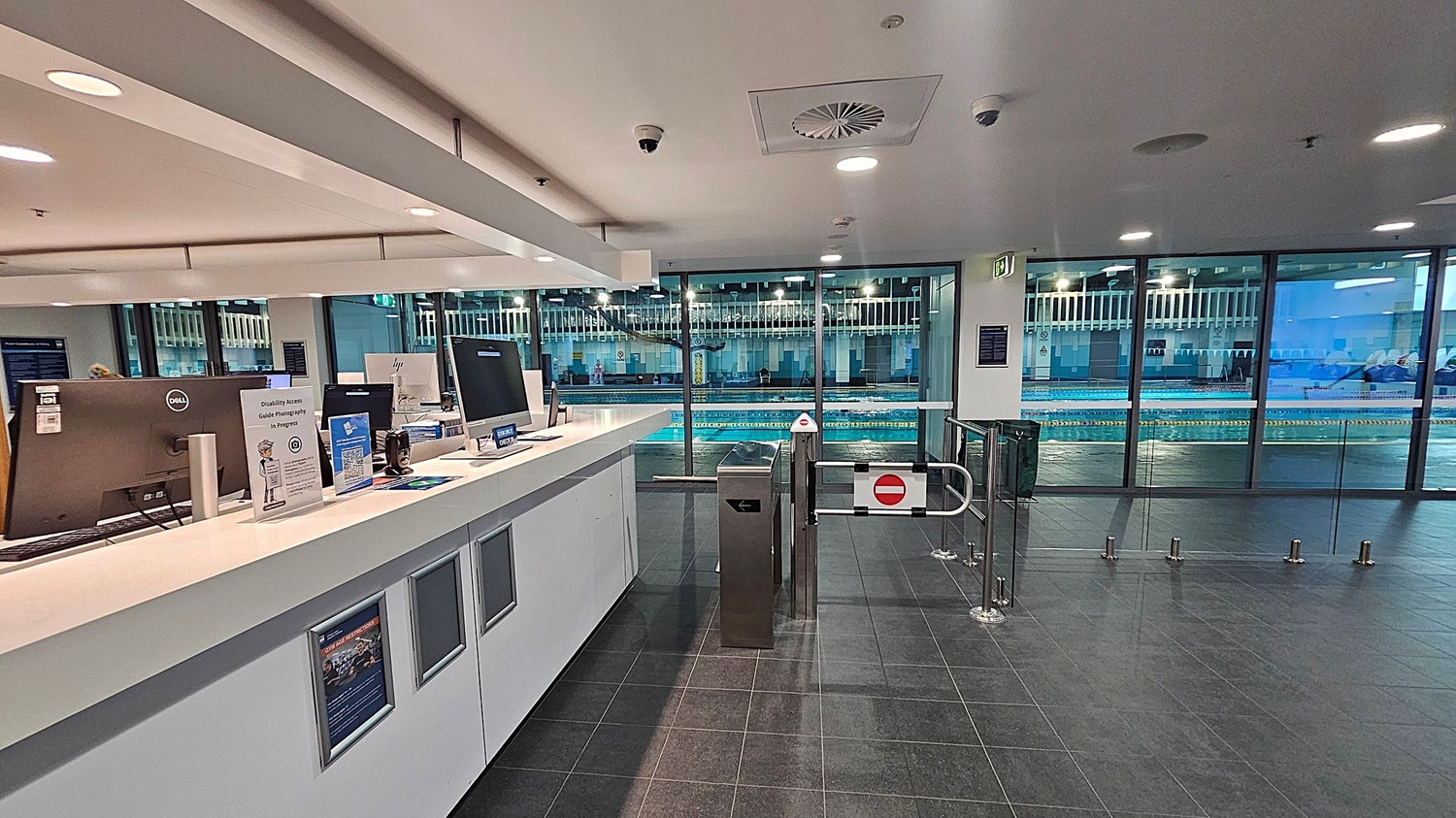 A modern indoor swimming pool facility with a reception area on the left featuring computer monitors, informational signs, and a counter. On the right, there is a turnstile gate with a "no entry" sign. Large glass windows in the background reveal a swimming pool with lanes and a few people swimming. The floor is dark grey tile, and the ceiling is white with recessed lighting and ventilation panels.