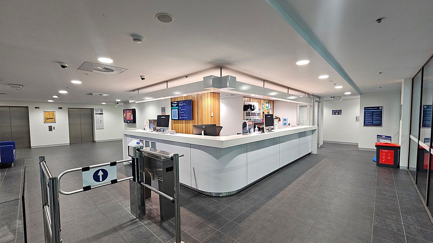A modern reception area with a large, white, L-shaped counter in the centre. Behind the counter are computer monitors and a wooden panelled wall with a digital screen. To the left, there are two closed lift doors and a blue seating area. On the right, a red bin and informational posters are visible. The floor is dark grey tile, and the ceiling is white with recessed lighting. A turnstile gate is in the foreground.