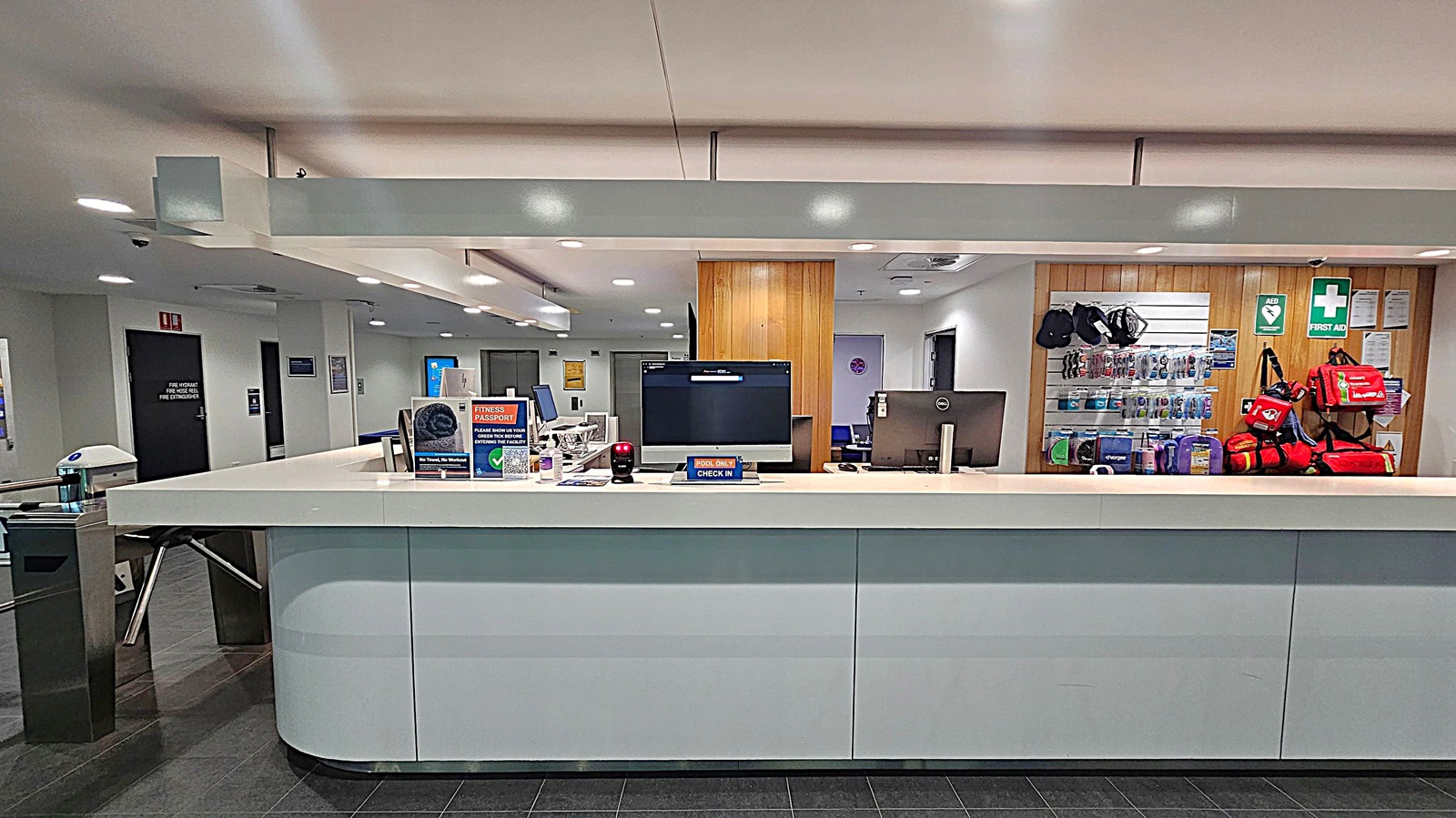 A reception desk with a white countertop and a computer monitor in the centre. On the left, there are brochures and a small electronic device. On the right, a wall-mounted display holds various first aid kits and equipment, with a green first aid sign above. The background features a wooden panel and a white wall with doors. The floor is dark grey tile, and the ceiling is white with recessed lighting.