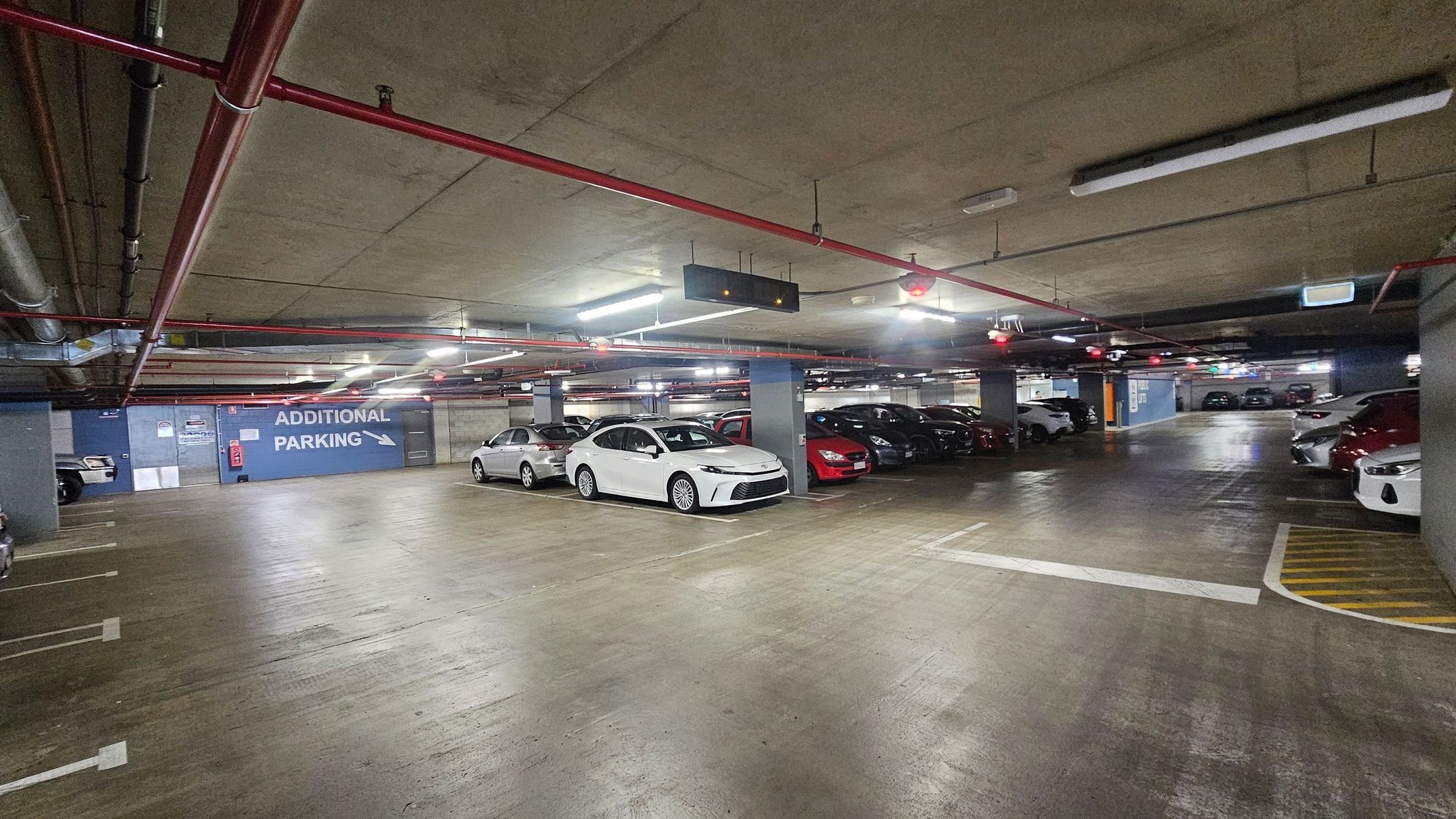 A spacious underground car park with concrete floors and ceiling, featuring several parked cars. A prominent blue sign on the left wall reads 'ADDITIONAL PARKING' with an arrow pointing right. Red and black pipes run along the ceiling, and fluorescent lights illuminate the area. Parking spaces are marked with white lines, and a digital sign hangs from the ceiling. The area feels enclosed with concrete pillars and walls.