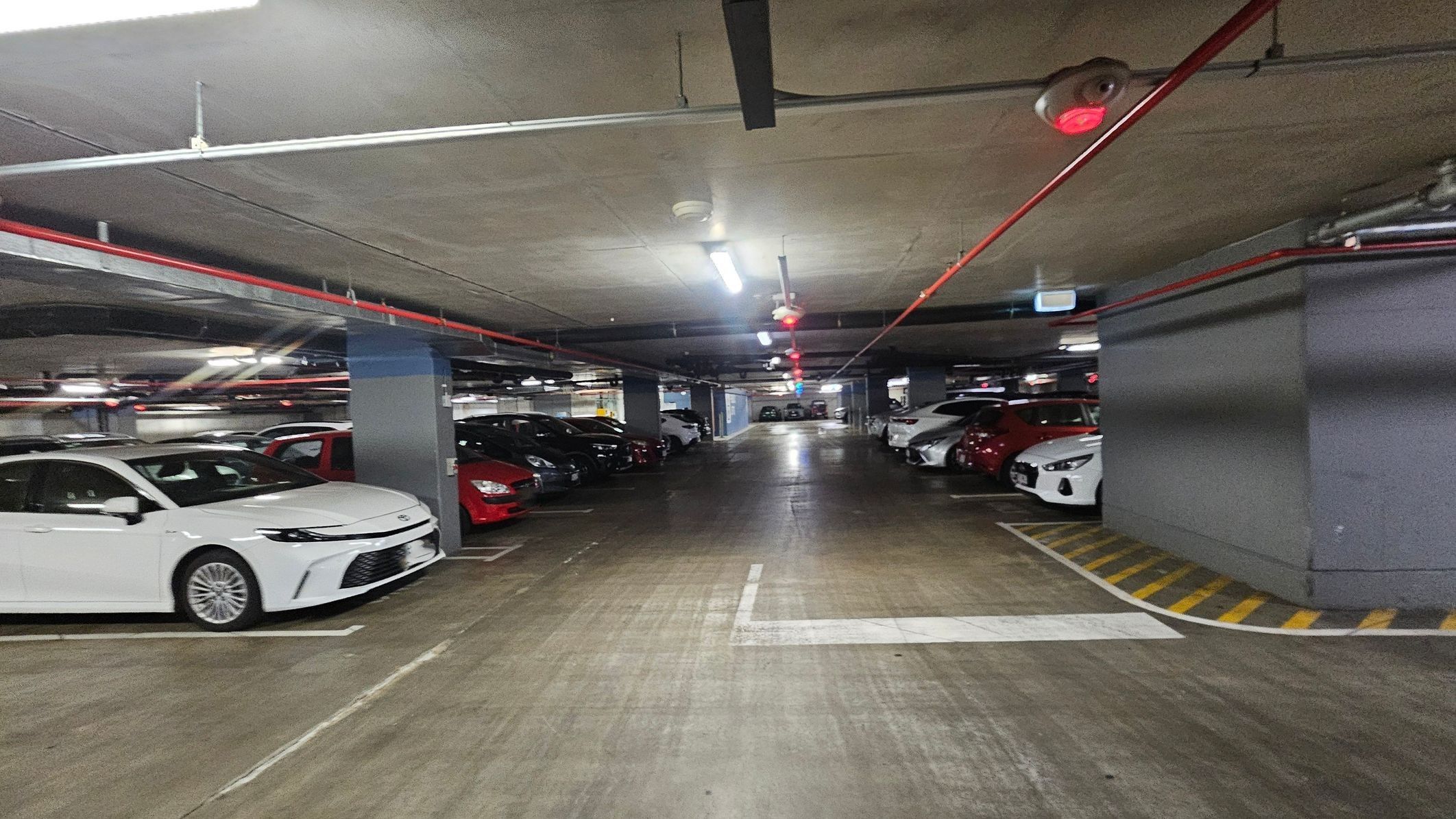 A parking garage with a large blue sign on the wall reading 'PUBLIC LIFTS' accompanied by lift symbols. The area is well-lit with fluorescent lights and has a concrete ceiling with visible pipes. The floor is a mix of worn concrete and painted sections. Several cars are parked in marked bays to the right. Payment machines are visible on the left, and a bollard stands near a yellow-striped area.