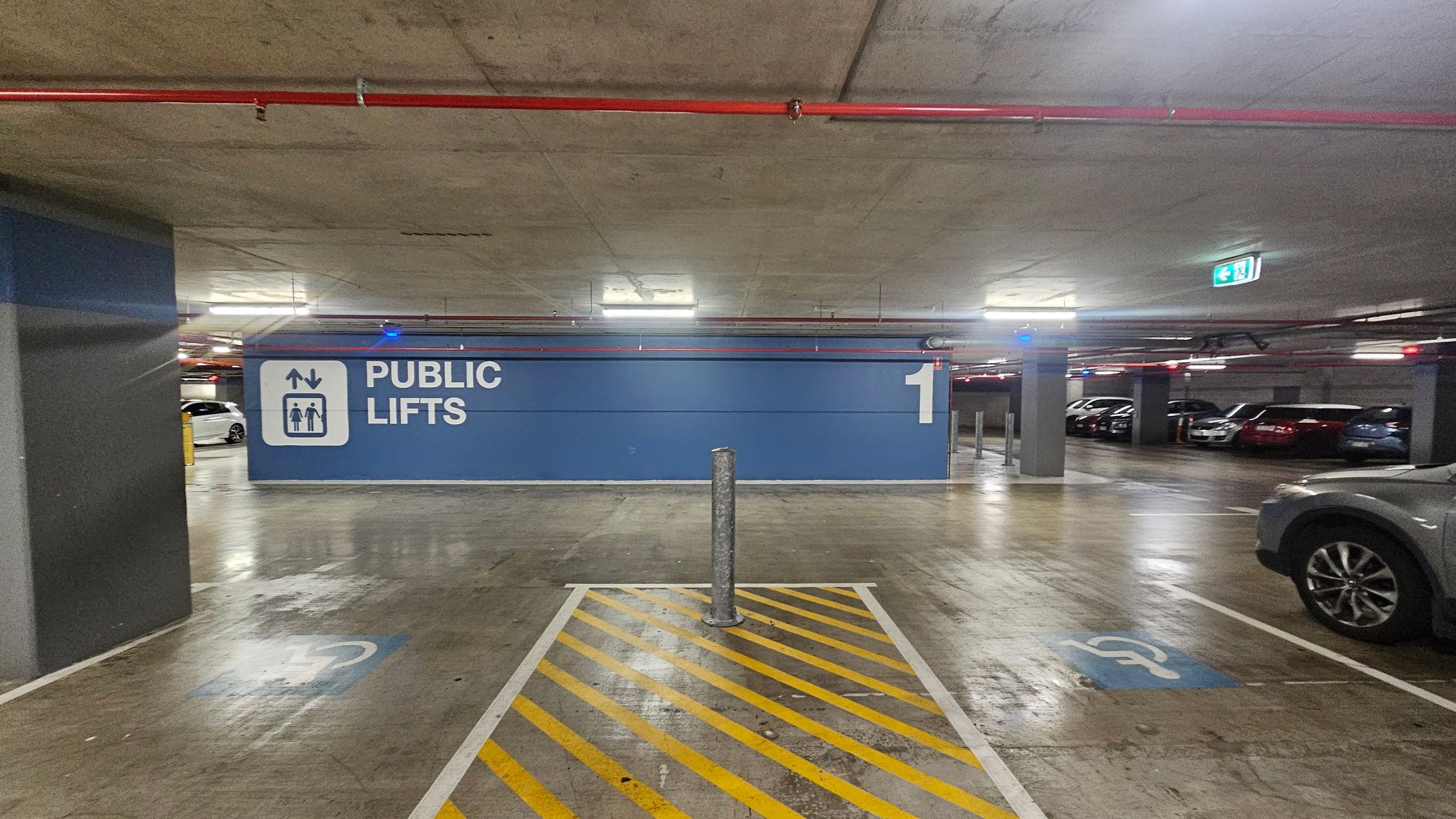 A parking garage with a large blue sign on the wall reading 'PUBLIC LIFTS' and the number '1'. The floor is concrete with yellow diagonal lines marking a no-parking zone, and a metal bollard in the centre. Several cars are parked to the right, and overhead, red pipes run along the ceiling. An illuminated exit sign is visible in the background.