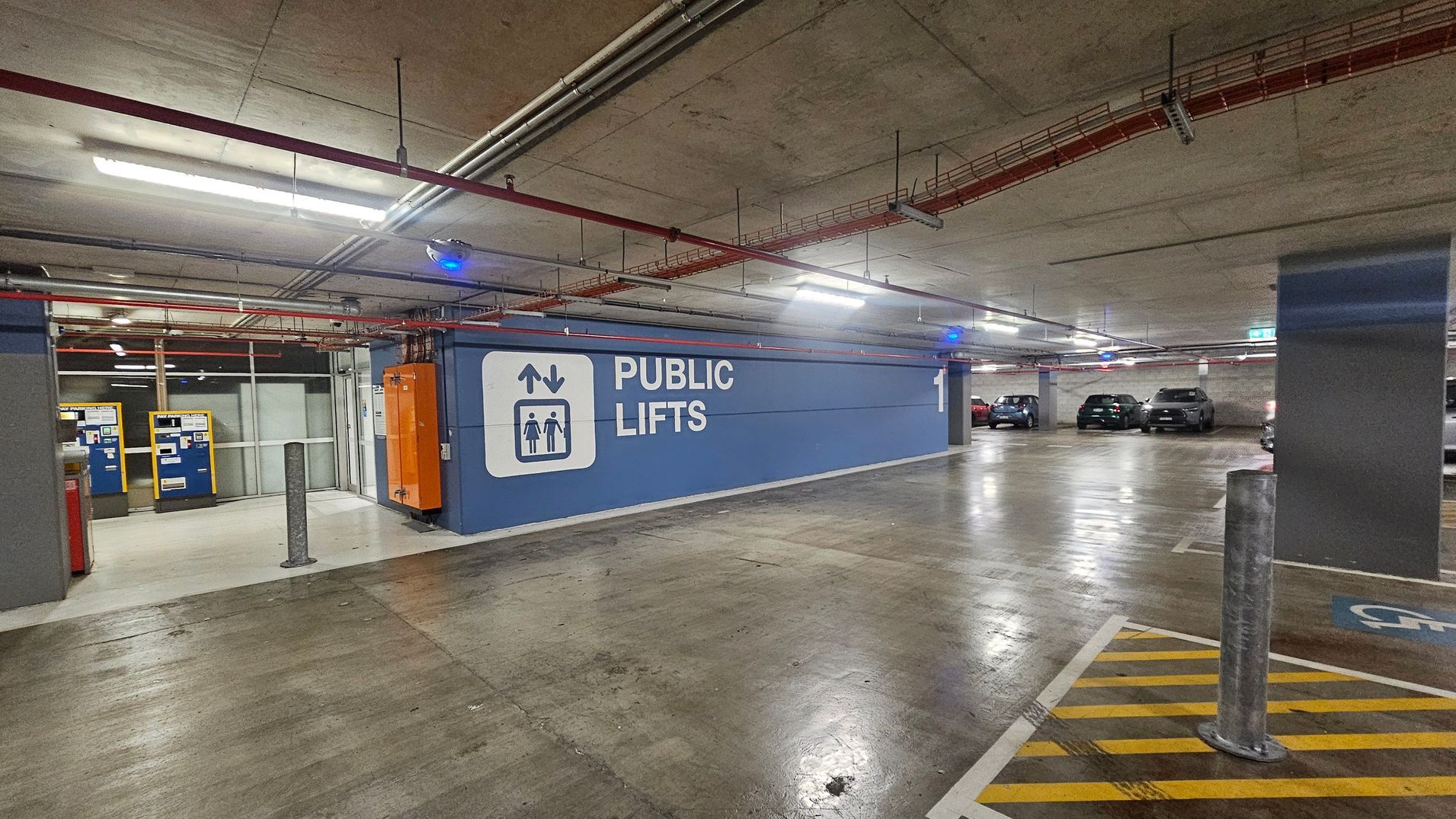 A spacious underground car park with several parked cars, including white and black vehicles. The floor is smooth concrete, and the ceiling is low with exposed pipes and fluorescent lighting. On the right, there are two yellow and blue payment machines next to a recycling bin station. A grey bollard stands nearby. The area feels enclosed with concrete pillars and walls, and there are clear parking space markings on the floor.