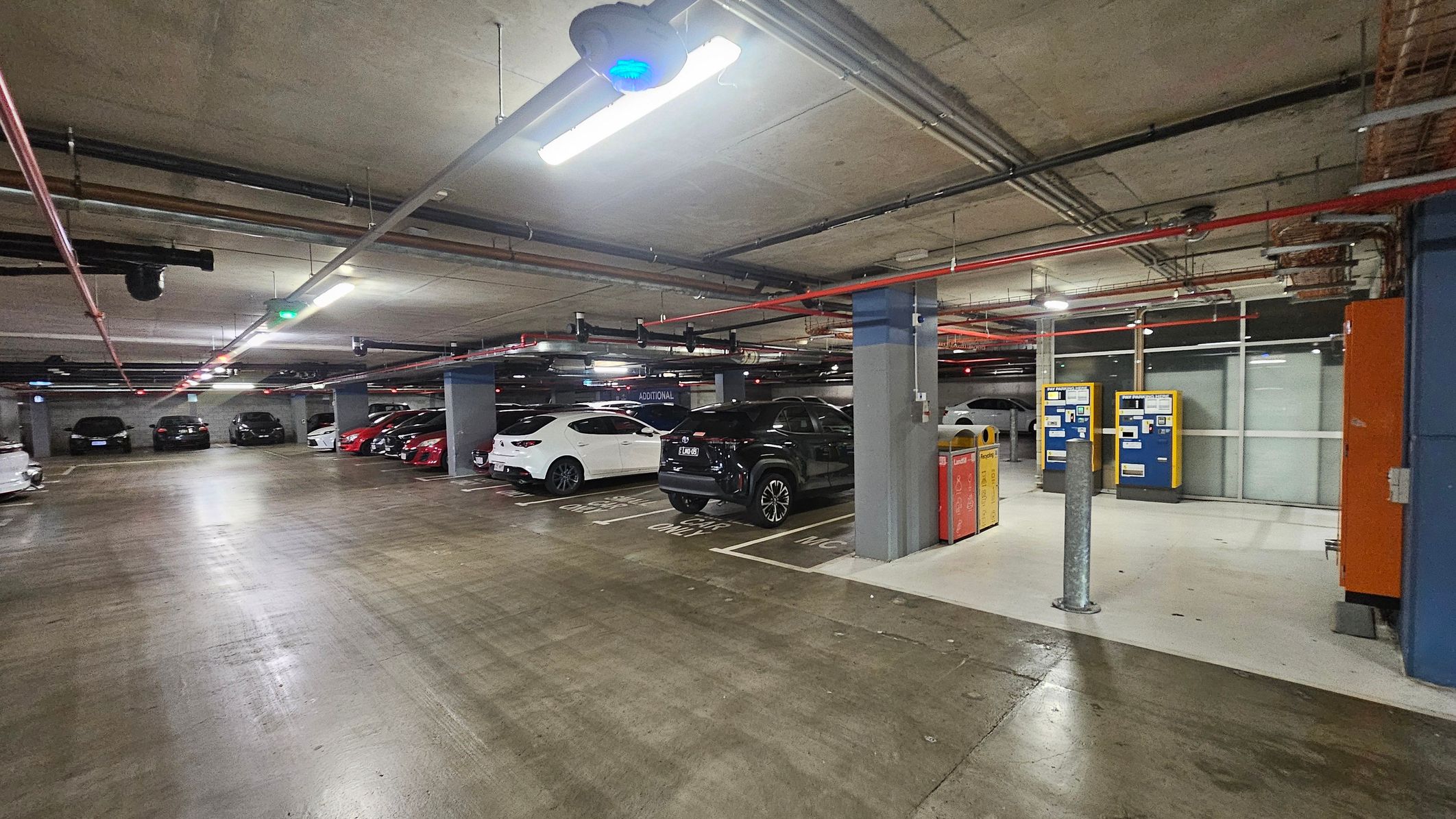 The image shows a parking garage with a focus on a glass entrance door on the left, leading to an interior space. Adjacent to the door is a blue wall with a mounted information board and an orange cabinet. The ceiling features exposed pipes and wiring. The floor is concrete, transitioning from a lighter to a darker shade. Several parked cars are visible in the background, separated by a metal railing and bollards.