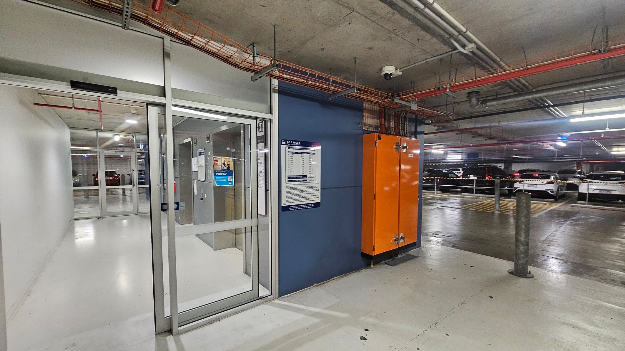 The image shows an indoor car park with a concrete ceiling and fluorescent lighting. On the left, a yellow pay parking machine is visible next to a glass door entrance leading to a corridor. An orange utility cabinet is mounted on a blue wall with signage. Several parked cars are seen on the right, separated by a metal railing. The floor transitions from light to dark grey, and a recycling bin is partially visible in the foreground.