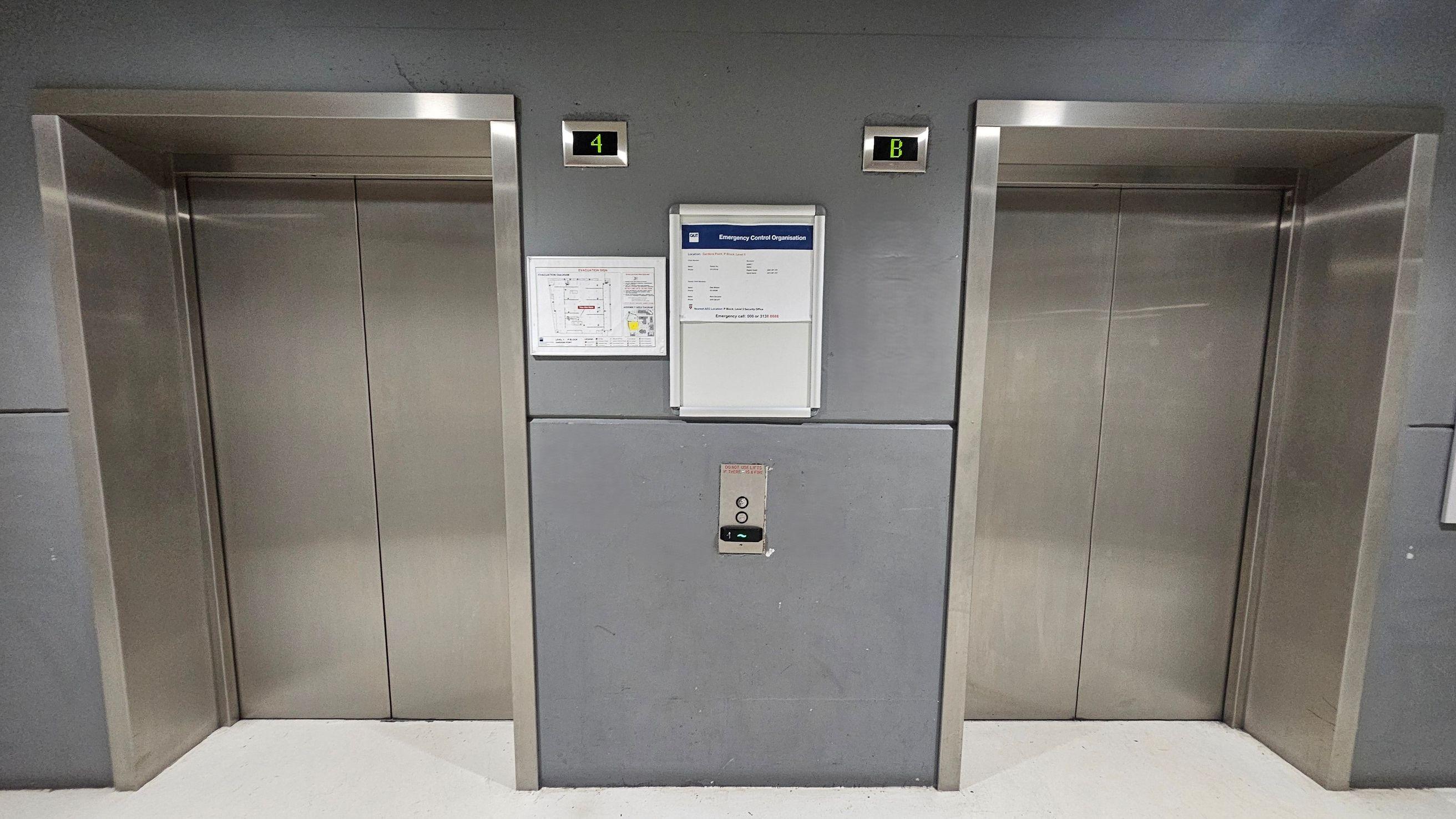The image shows two closed lift doors set into a grey wall, each framed with stainless steel. Above the left lift is a digital display showing the number 4, and above the right lift is a display showing the letter B. Between the lifts, there is a control panel with two buttons and a small display. A notice board with emergency contact information and a floor plan is mounted on the wall. The floor is a smooth, light-coloured surface.