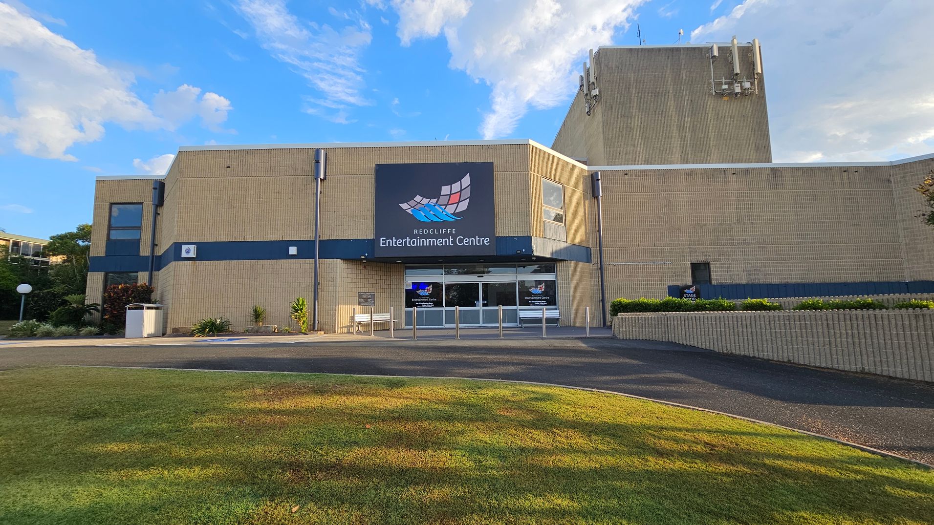 A beige brick building with a large sign reading "Redcliffe Entertainment Centre" above the entrance. The entrance has glass doors and is flanked by small plants and a bench on the left. The building has a blue horizontal stripe and several small windows. In the foreground, there is a grassy area leading to a paved path. The sky is visible above the building. A beige brick building with a large sign reading "Redcliffe Entertainment Centre" above the entrance. The entrance has glass doors and is flanked by small plants and a bench on the left. The building has a blue horizontal stripe and several small windows. In the foreground, there is a grassy area leading to a paved path. The sky is visible above the building.