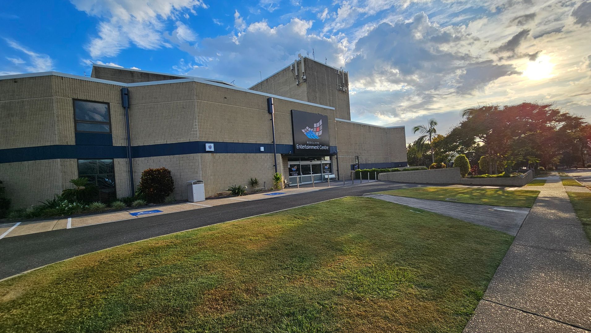 A large building with a beige exterior and a sign reading "Entertainment Centre" above the entrance. The entrance has glass doors and is flanked by small plants. To the right, a well-maintained lawn and a path lead to a garden area with trees and shrubs. The sun is low in the sky, casting long shadows on the ground. The sky is partly cloudy, and the overall setting appears calm and organised. A large building with a beige exterior and a sign reading "Entertainment Centre" above the entrance. The entrance has glass doors and is flanked by small plants. To the right, a well-maintained lawn and a path lead to a garden area with trees and shrubs. The sun is low in the sky, casting long shadows on the ground. The sky is partly cloudy, and the overall setting appears calm and organised.