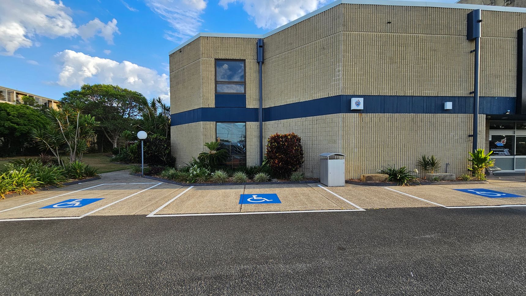 A building with light-coloured brick walls and a dark blue horizontal stripe. In front, there are three parking spaces marked with blue disabled signs. A small garden with various plants and a white spherical lamp post is on the left. A rubbish bin is positioned near the building. The entrance to the building is on the right, with glass doors and a sign. The ground is a mix of asphalt and concrete. A building with light-coloured brick walls and a dark blue horizontal stripe. In front, there are three parking spaces marked with blue disabled signs. A small garden with various plants and a white spherical lamp post is on the left. A rubbish bin is positioned near the building. The entrance to the building is on the right, with glass doors and a sign. The ground is a mix of asphalt and concrete.