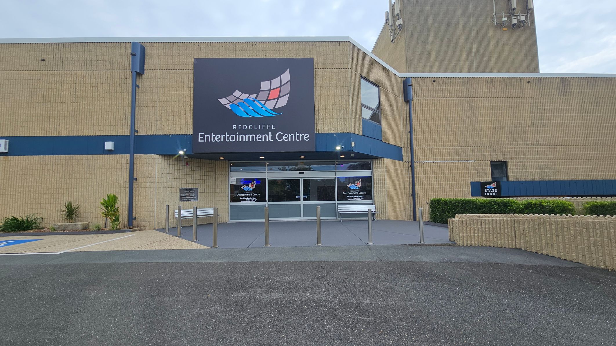 A brick building with a large sign reading "Redcliffe Entertainment Centre" above the entrance. The entrance has glass doors and is flanked by two white benches. There are several metal bollards in front of the entrance. To the left, there are small plants and a designated parking area. On the right, a sign indicates a "Stage Door" near a hedge. The ground is paved with asphalt and concrete, and the building has a blue trim. A brick building with a large sign reading "Redcliffe Entertainment Centre" above the entrance. The entrance has glass doors and is flanked by two white benches. There are several metal bollards in front of the entrance. To the left, there are small plants and a designated parking area. On the right, a sign indicates a "Stage Door" near a hedge. The ground is paved with asphalt and concrete, and the building has a blue trim.