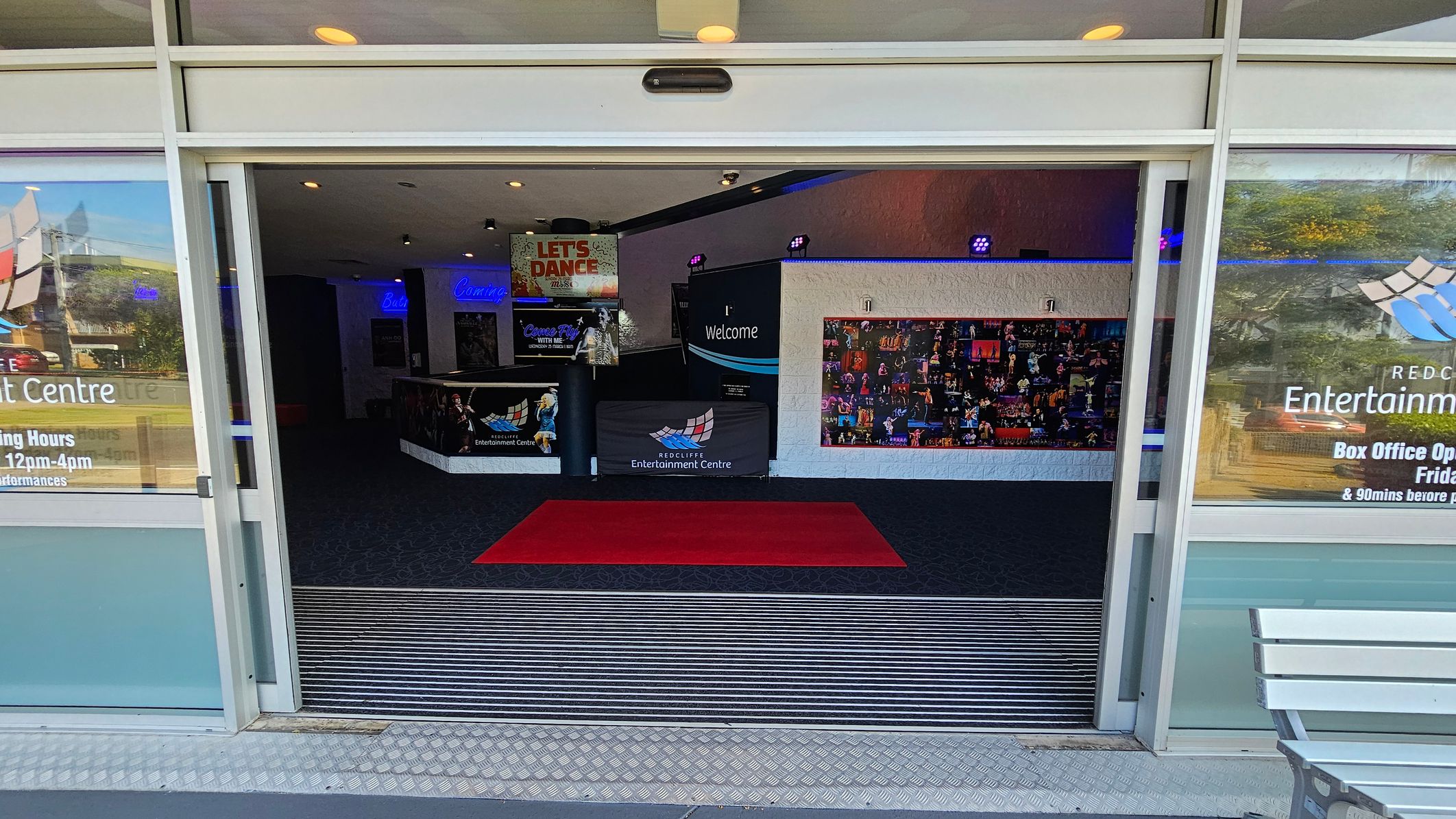 Entrance to a building with glass doors open, revealing an interior with a red carpet on a dark floor. The left and right sides have posters and signage, including "Redcliffe Entertainment Centre" and box office hours. Inside, a "Welcome" sign and a collage of images are visible on the walls. The ceiling has recessed lighting, and a bench is partially visible on the right outside. Entrance to a building with glass doors open, revealing an interior with a red carpet on a dark floor. The left and right sides have posters and signage, including "Redcliffe Entertainment Centre" and box office hours. Inside, a "Welcome" sign and a collage of images are visible on the walls. The ceiling has recessed lighting, and a bench is partially visible on the right outside.