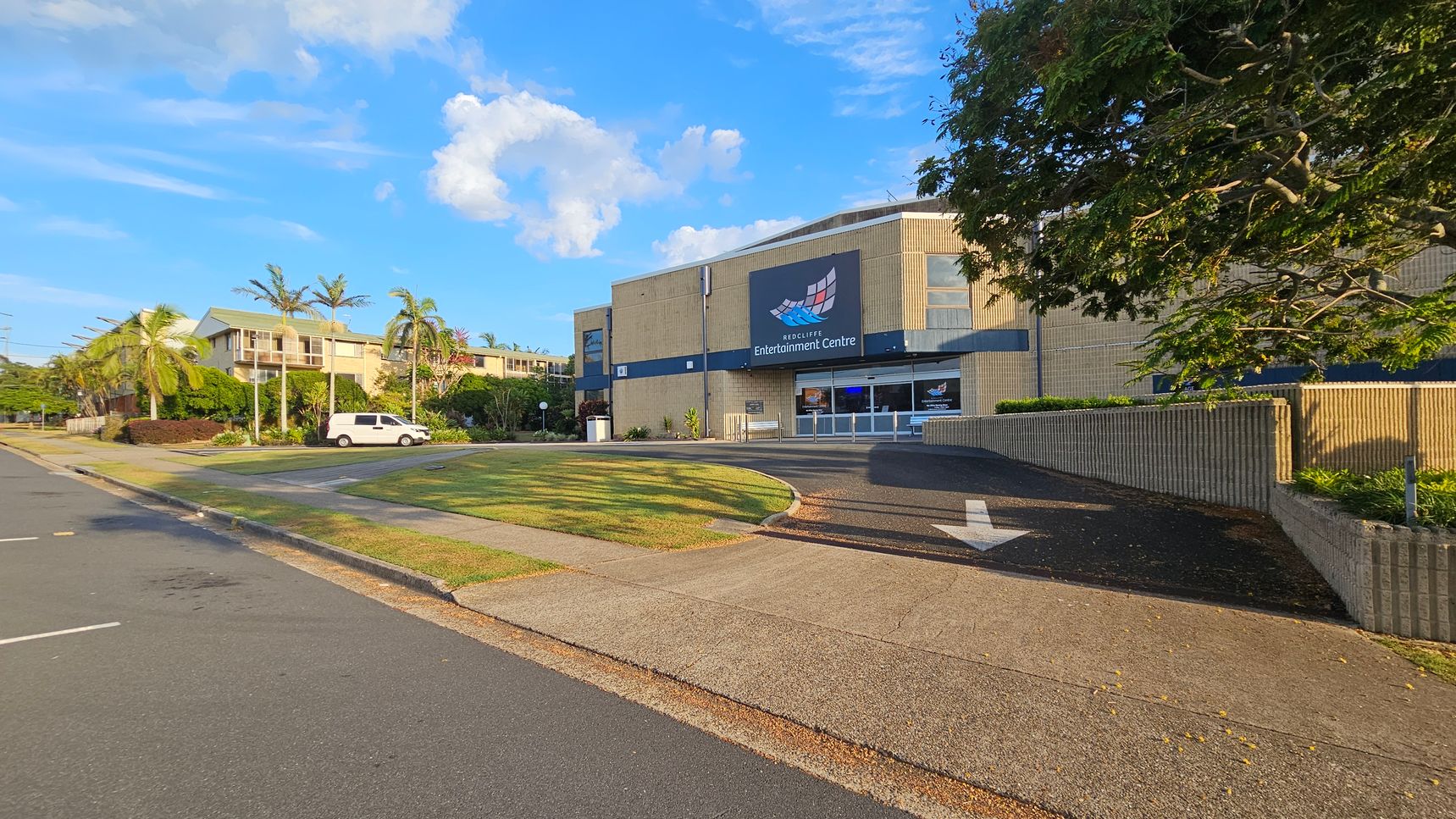 A street view with a large building on the right labelled "Redcliffe Entertainment Centre". The building is beige with a dark blue sign. A tree with green leaves partially shades the building. On the left, a white van is parked near a row of palm trees and residential buildings. The foreground features a paved footpath and a grassy area, with a road running alongside. The sky is clear with a few clouds. A street view with a large building on the right labelled "Redcliffe Entertainment Centre". The building is beige with a dark blue sign. A tree with green leaves partially shades the building. On the left, a white van is parked near a row of palm trees and residential buildings. The foreground features a paved footpath and a grassy area, with a road running alongside. The sky is clear with a few clouds.