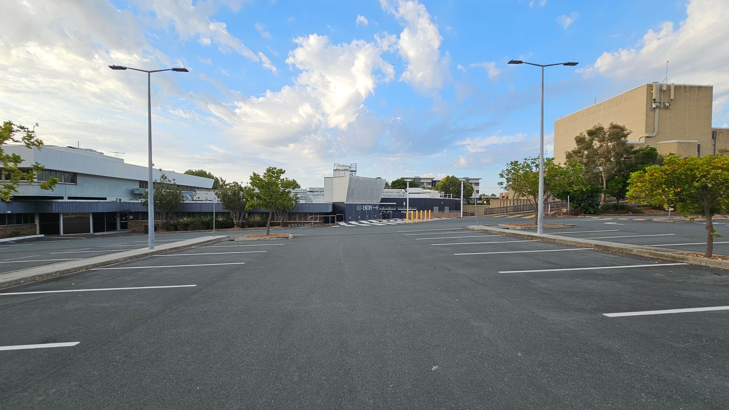 An empty car park with marked spaces and two tall streetlights. On the left, a modern building with a flat roof and large windows. In the centre, a structure with a sign indicating "Entry" and a pedestrian crossing. On the right, a large beige building with trees and shrubs around it. The ground is asphalt, and there are small trees planted along the edges. An empty car park with marked spaces and two tall streetlights. On the left, a modern building with a flat roof and large windows. In the centre, a structure with a sign indicating "Entry" and a pedestrian crossing. On the right, a large beige building with trees and shrubs around it. The ground is asphalt, and there are small trees planted along the edges.