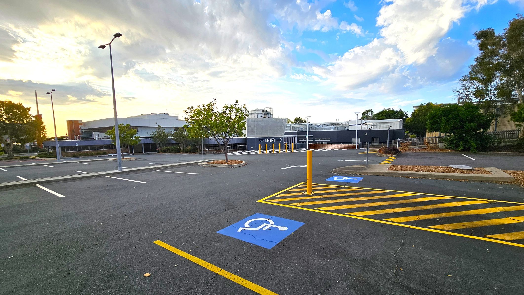 An empty car park with marked parking spaces, including a blue disabled parking sign on the ground. Yellow-striped lines are visible near the foreground. Several tall streetlights are scattered throughout the area. In the background, there is a large building with a sign indicating "ENTRY" and some trees and shrubs around the perimeter. The sky is partly cloudy with a mix of blue and light hues. An empty car park with marked parking spaces, including a blue disabled parking sign on the ground. Yellow-striped lines are visible near the foreground. Several tall streetlights are scattered throughout the area. In the background, there is a large building with a sign indicating "ENTRY" and some trees and shrubs around the perimeter. The sky is partly cloudy with a mix of blue and light hues.