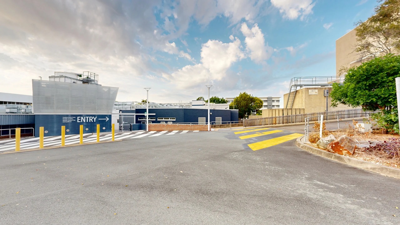 A paved area with a marked pedestrian crossing in yellow and white lines. On the left, a blue building with "ENTRY" and an arrow is visible, along with several yellow bollards. In the background, there are trees and additional buildings with metal railings. On the right, a small landscaped area with rocks and shrubs is present. The sky is partly cloudy. A paved area with a marked pedestrian crossing in yellow and white lines. On the left, a blue building with "ENTRY" and an arrow is visible, along with several yellow bollards. In the background, there are trees and additional buildings with metal railings. On the right, a small landscaped area with rocks and shrubs is present. The sky is partly cloudy.