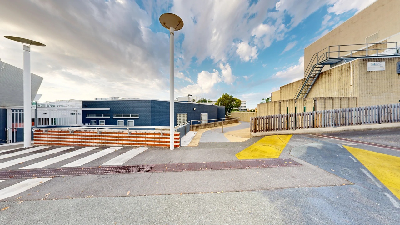A paved area with a zebra crossing in the foreground. On the left, there is a blue building with white-framed windows and a red and white striped barrier. Two tall street lamps are visible. On the right, a beige building with a metal staircase and a wooden fence. The background features a tree and some distant buildings. The ground has yellow painted markings, and the sky is partly cloudy. A paved area with a zebra crossing in the foreground. On the left, there is a blue building with white-framed windows and a red and white striped barrier. Two tall street lamps are visible. On the right, a beige building with a metal staircase and a wooden fence. The background features a tree and some distant buildings. The ground has yellow painted markings, and the sky is partly cloudy.