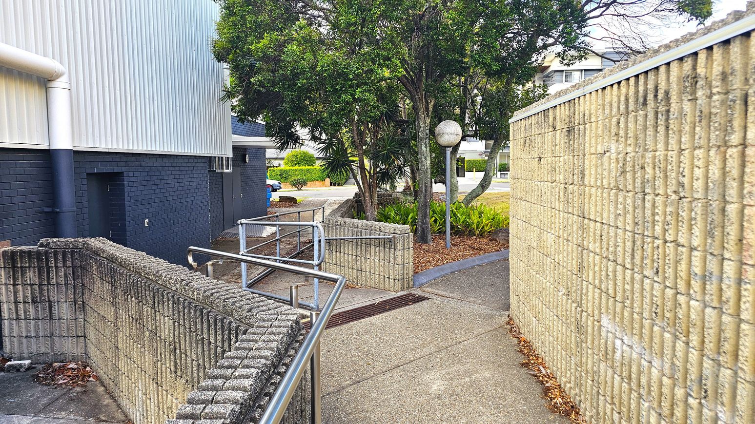 A narrow pathway with a textured concrete wall on the right and a metal railing on the left. The ground is paved with concrete. In the middle, there are several trees and a spherical streetlamp. The background shows a building with a corrugated metal exterior and a dark brick wall. Some greenery and a parked car are visible further back. A narrow pathway with a textured concrete wall on the right and a metal railing on the left. The ground is paved with concrete. In the middle, there are several trees and a spherical streetlamp. The background shows a building with a corrugated metal exterior and a dark brick wall. Some greenery and a parked car are visible further back.