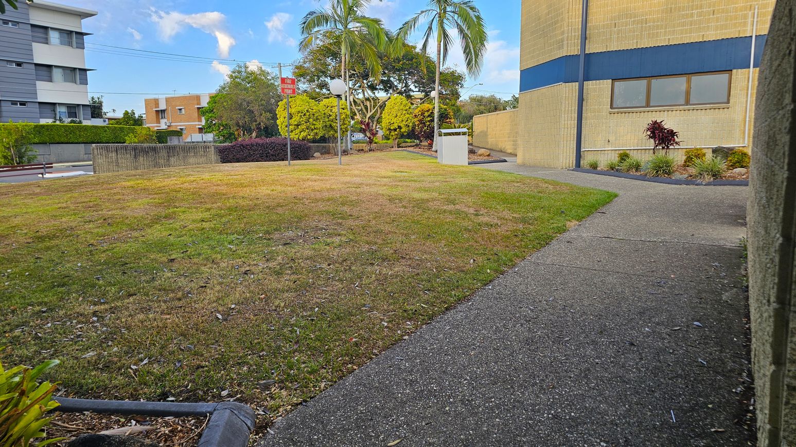 A grassy area with patches of brown is in the foreground, bordered by a concrete path on the right. In the background, there are several trees, including palm trees, and a few buildings. A signpost and a white structure are visible near the centre. The building on the right has a beige and blue exterior with windows, while another building on the left is grey and white. Bushes and small plants are along the path. A grassy area with patches of brown is in the foreground, bordered by a concrete path on the right. In the background, there are several trees, including palm trees, and a few buildings. A signpost and a white structure are visible near the centre. The building on the right has a beige and blue exterior with windows, while another building on the left is grey and white. Bushes and small plants are along the path.