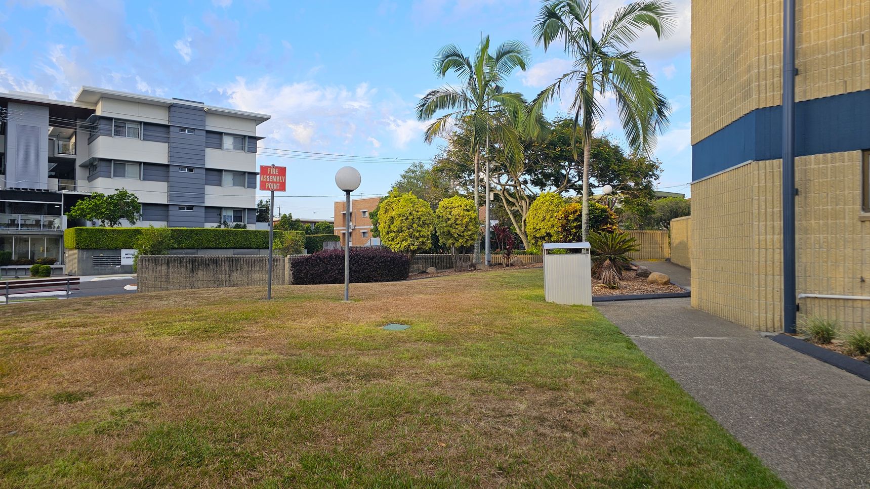A grassy area with patches of brown is in the foreground. On the left, a modern building with grey and white horizontal panels is visible. A "Fire Assembly Point" sign and a spherical streetlamp are near the centre. On the right, a beige building with a blue stripe is partially visible. Several palm trees and bushes are in the background, along with a small pathway and a bin. A low brick wall separates the grassy area from the road. A grassy area with patches of brown is in the foreground. On the left, a modern building with grey and white horizontal panels is visible. A "Fire Assembly Point" sign and a spherical streetlamp are near the centre. On the right, a beige building with a blue stripe is partially visible. Several palm trees and bushes are in the background, along with a small pathway and a bin. A low brick wall separates the grassy area from the road.